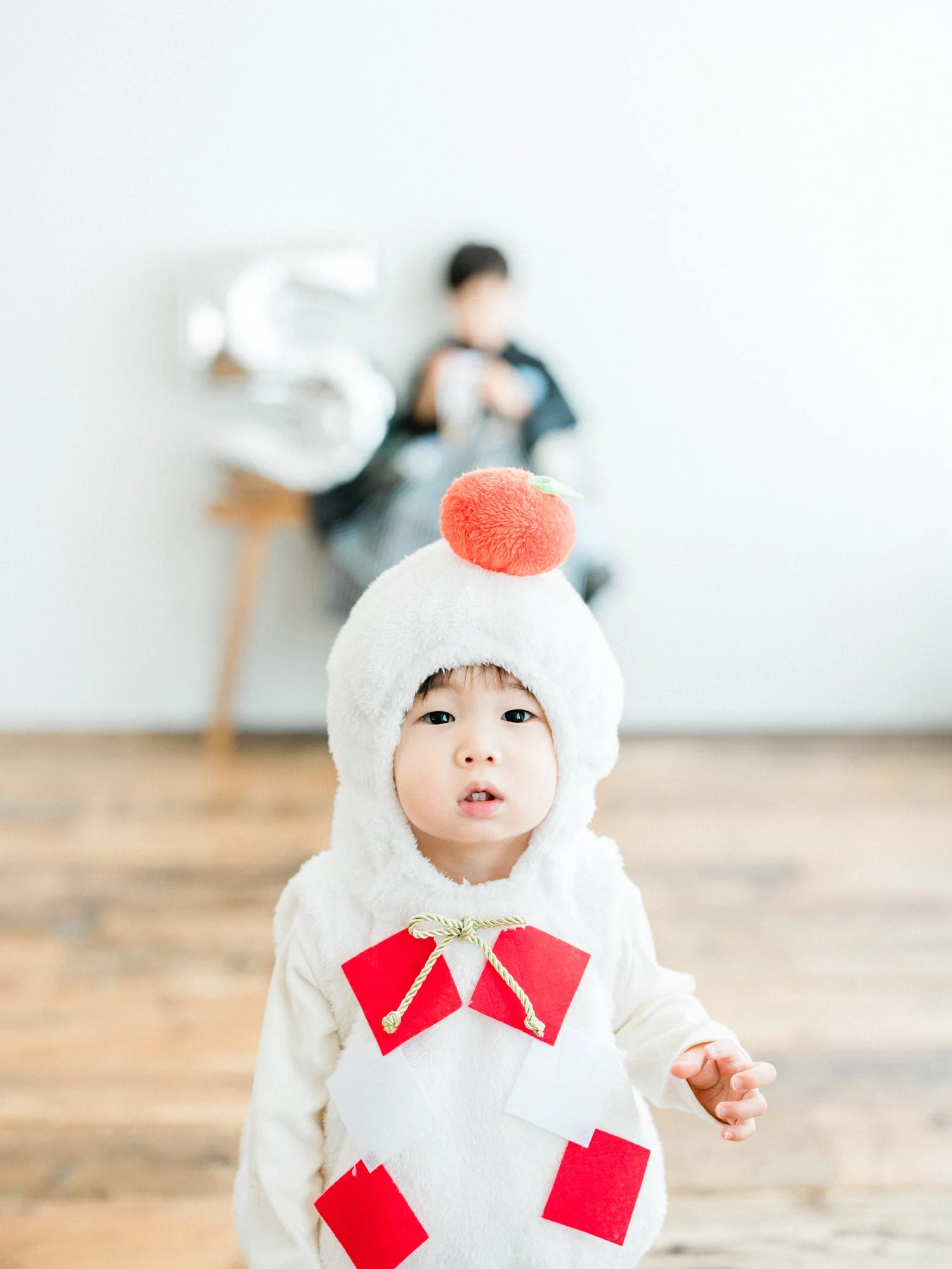 子供が赤い折り紙の飾りと和柄の飾りをつけた、お餅のコスチュームを着ている。背景はシンプルな白壁と木の床。