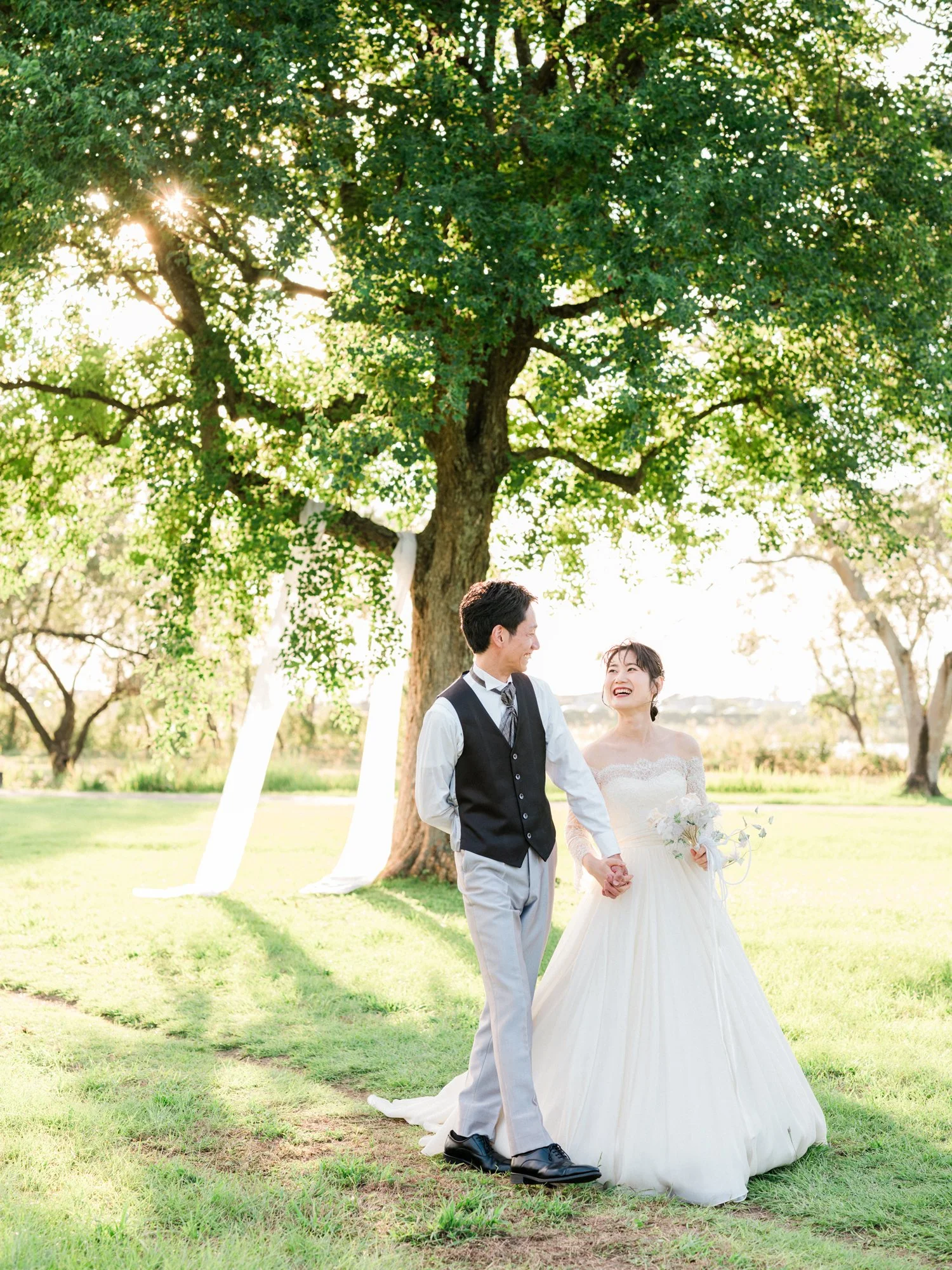 緑豊かな公園の木の下で結婚式を挙げている新郎新婦。新婦は白いドレスを着て花束を持ち、笑顔で旦那さんと見つめ合っている。背景には大きな木と太陽の光が差し込む風景。