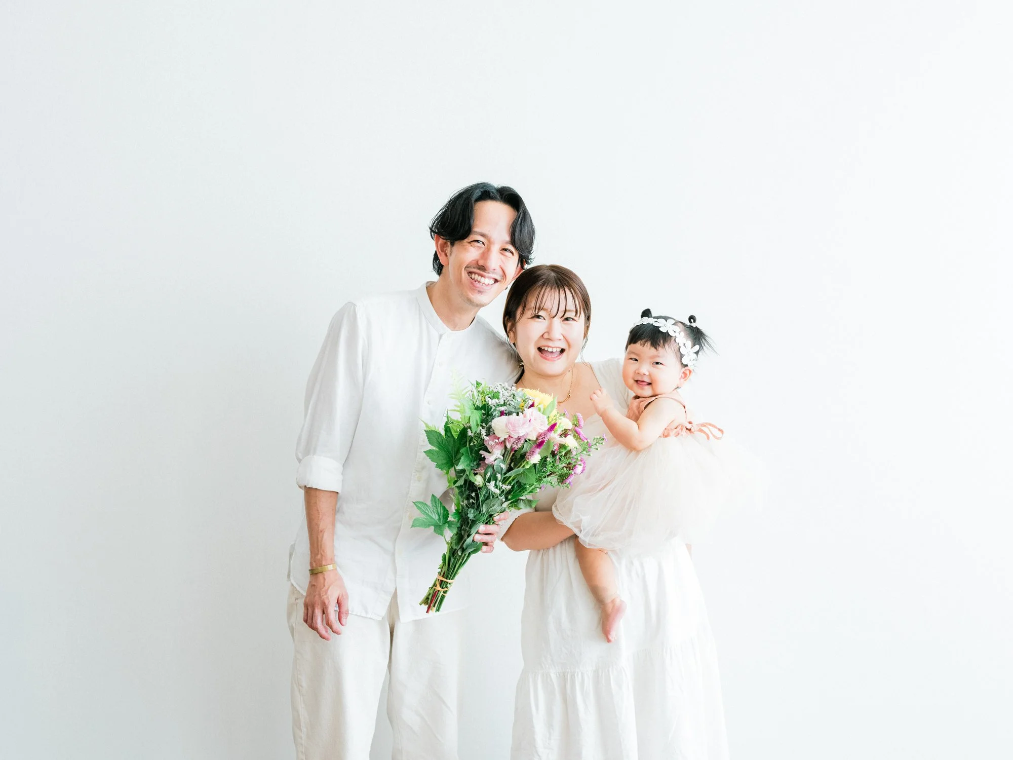 白い服を着た男性と女性、子供が花束を持ち笑顔で写っている。
