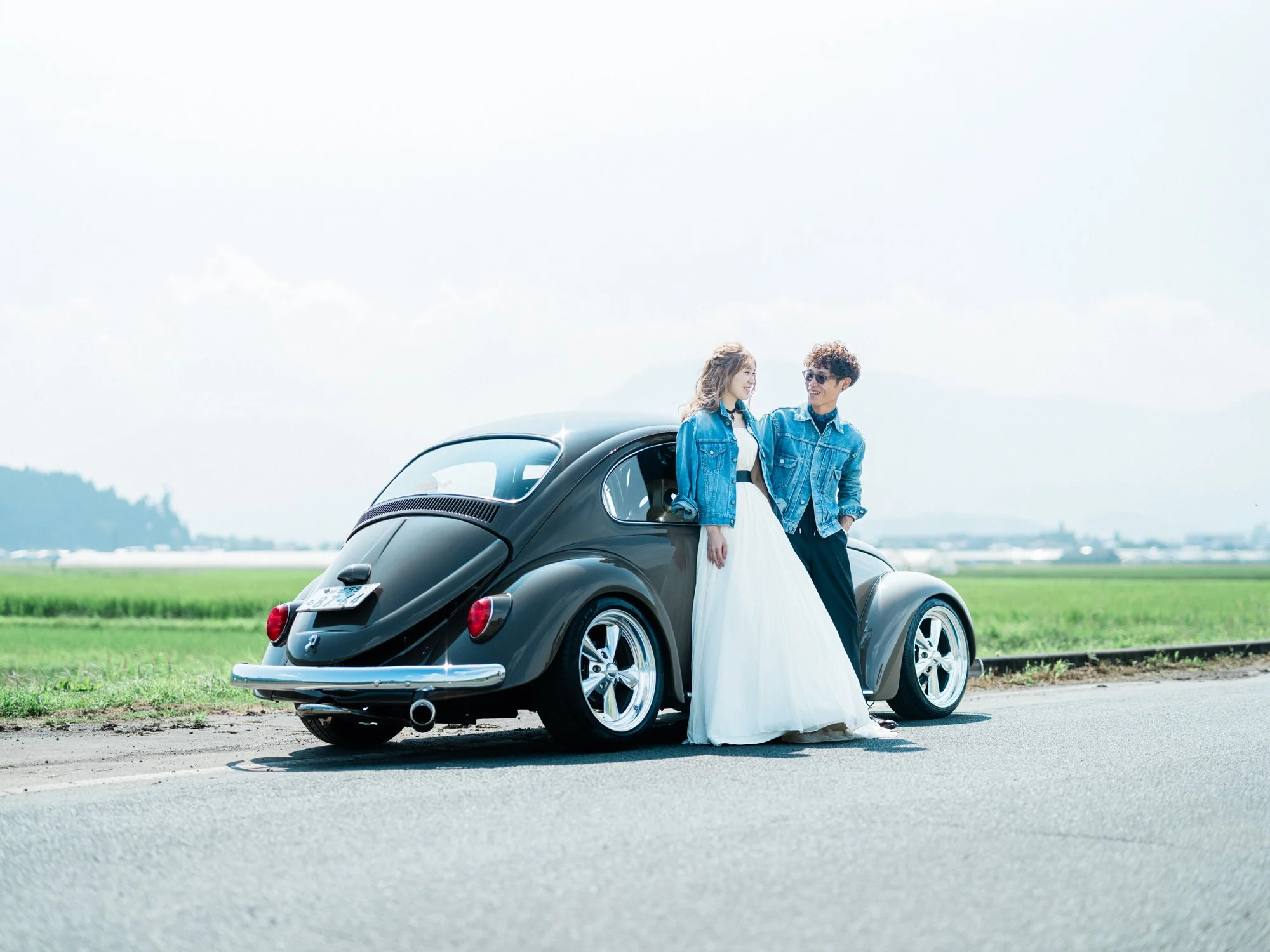空の広場で、車の横に立つ男性と女性のカップル。女性は白いドレスとデニムジャケットを着ており、男性はデニムジャケットを着ている。背景は青空と遠くの山々。