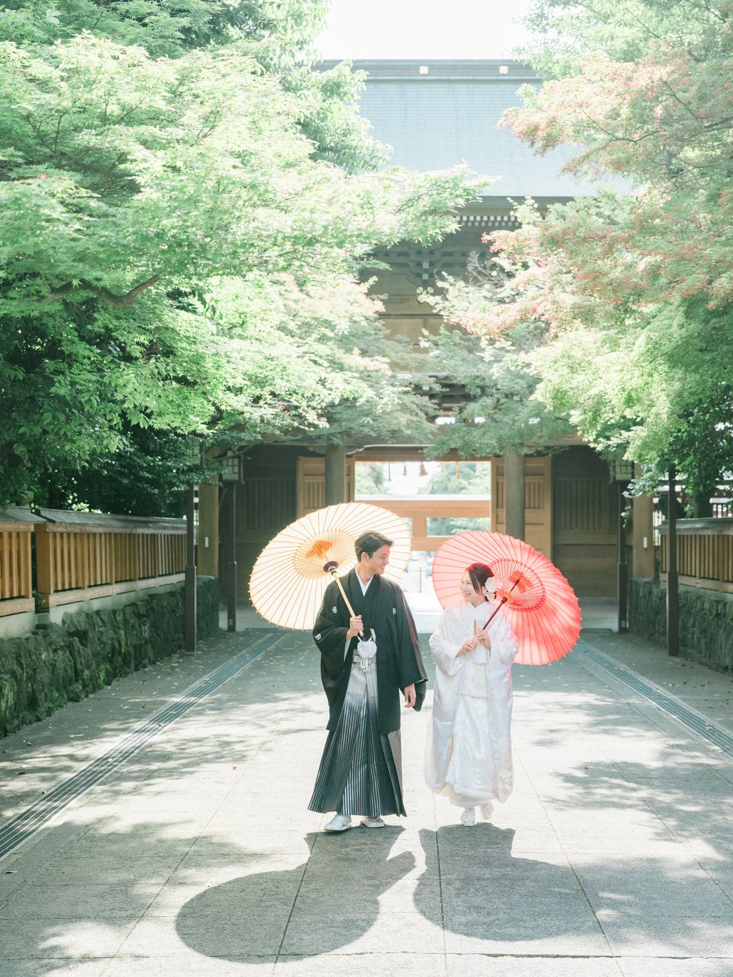 神社の鳥居の前で和装の新郎新婦が微笑みながらおしゃべりし、傘をさしている。背景には緑豊かな木々と門が見える。