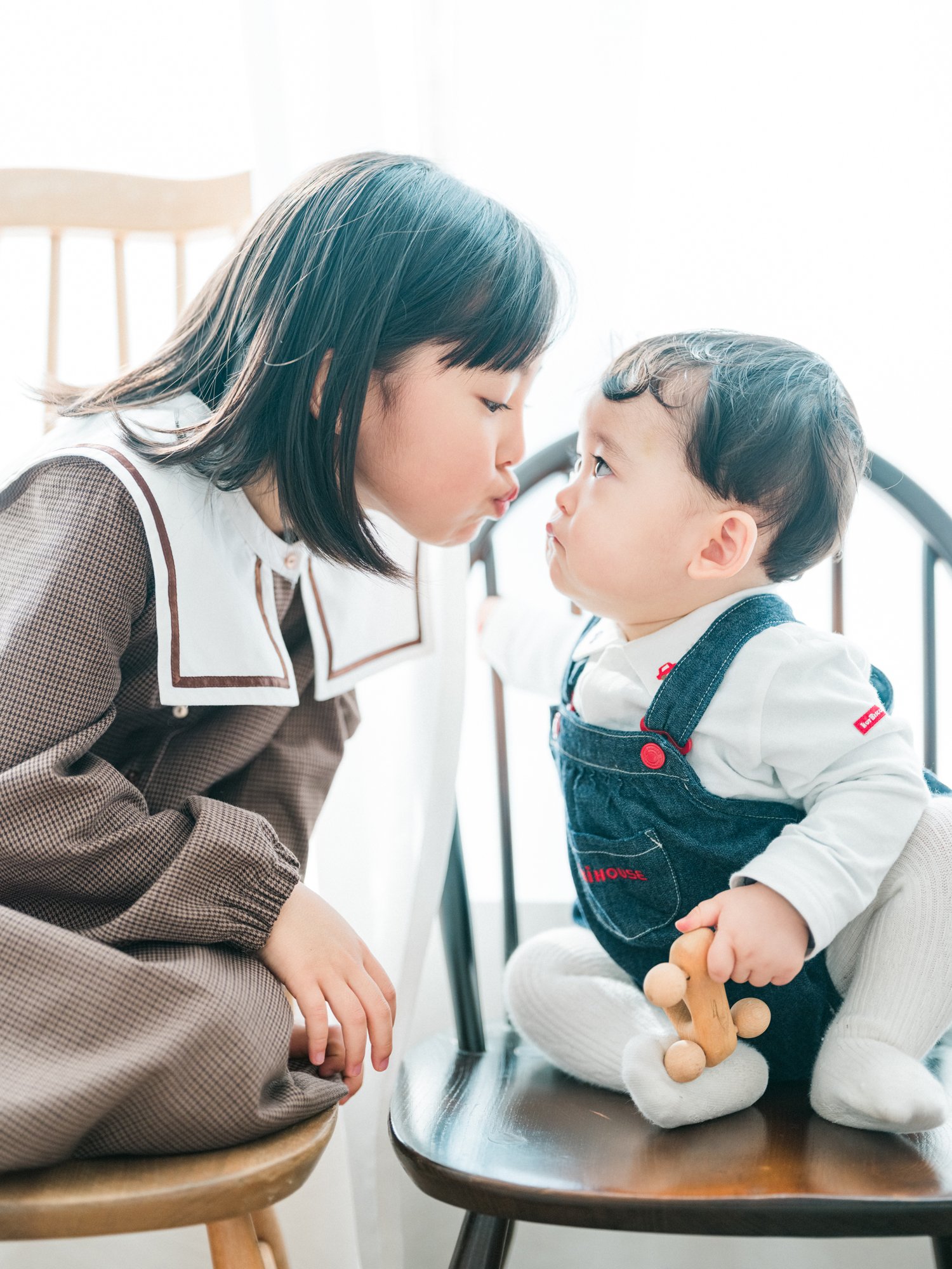 お姉ちゃんと幼い男の子がテーブル越しにおでこをつき合わせている。背景は明るく、女性はブラウンの格子柄の服、男の子は白いシャツにデニムのオーバーオールを着て、木製のおもちゃを手に持っている。