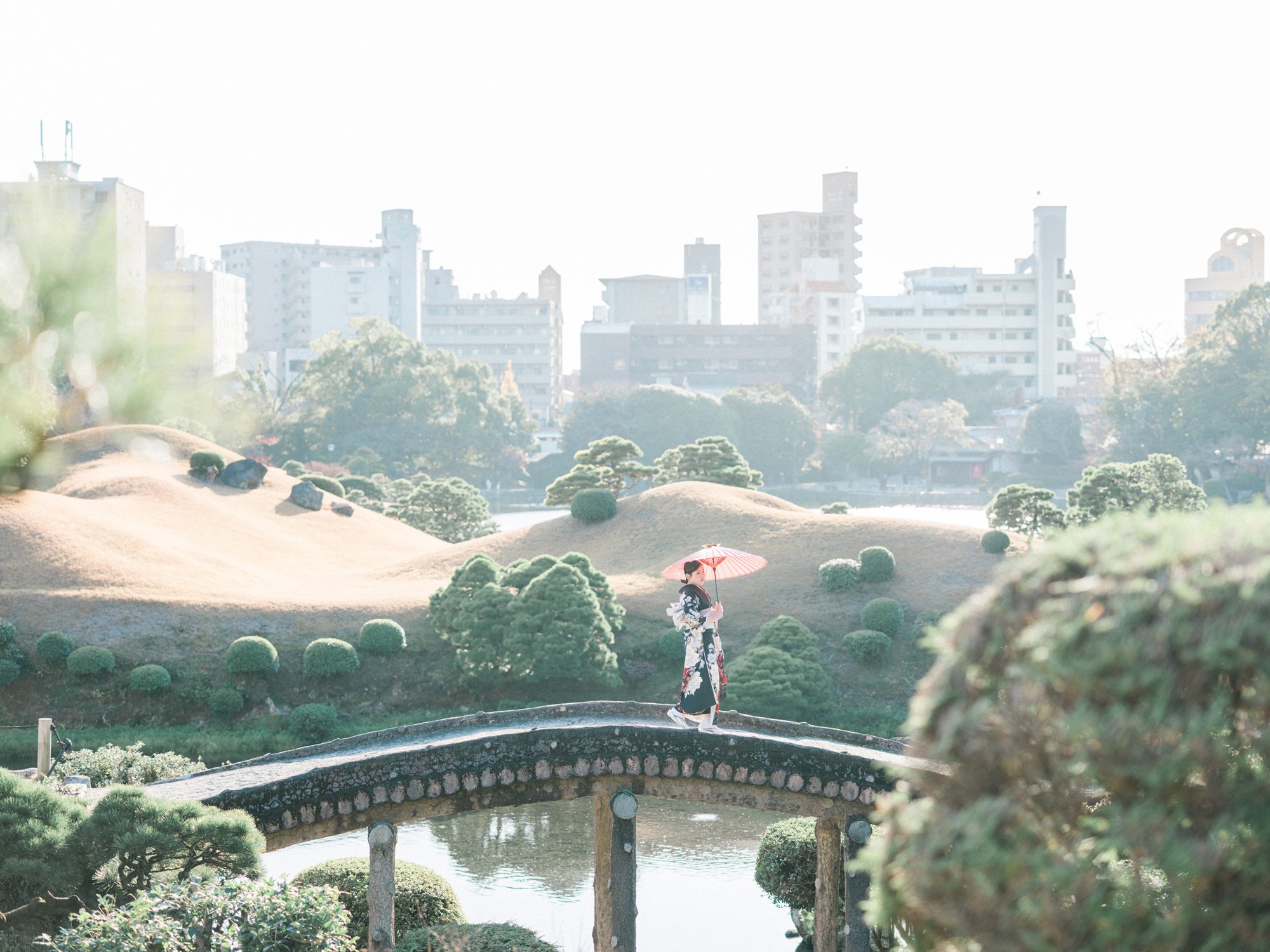 都市の中の日本庭園の風景で、女性が着物を着て傘をさし、橋を渡っている。背景にビル群が見える。