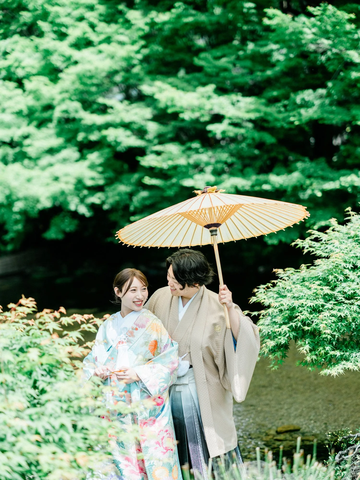 男性と女性が日本の伝統衣装を着て水前寺公園で微笑み合いながら歩いており、男性は和傘を持っている。背景に緑の木々と池がある風景です。