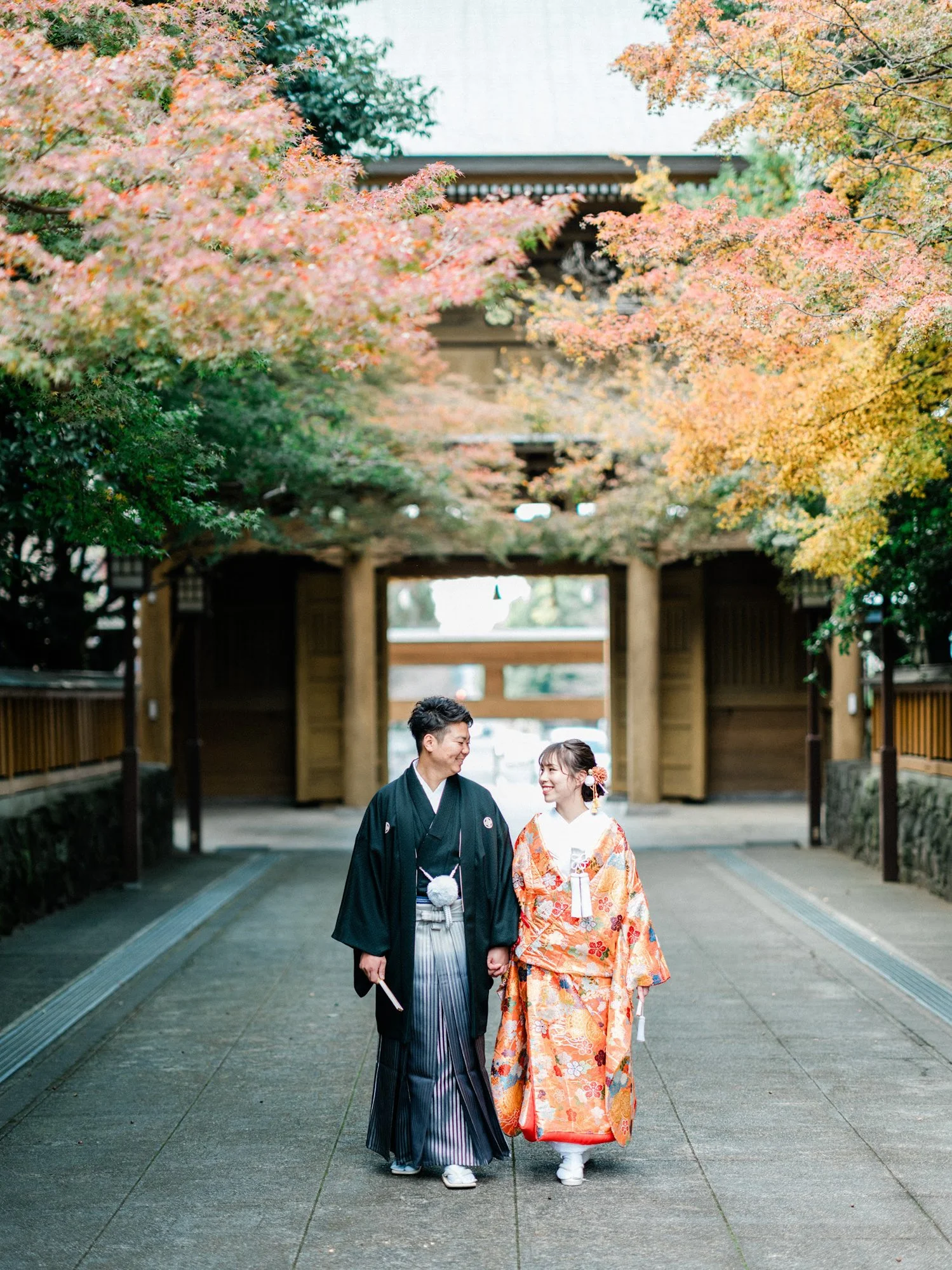 和装を着た夫婦が手をつないで歩いている。背景には紅葉した木々と神社の鳥居と入口門が見える。