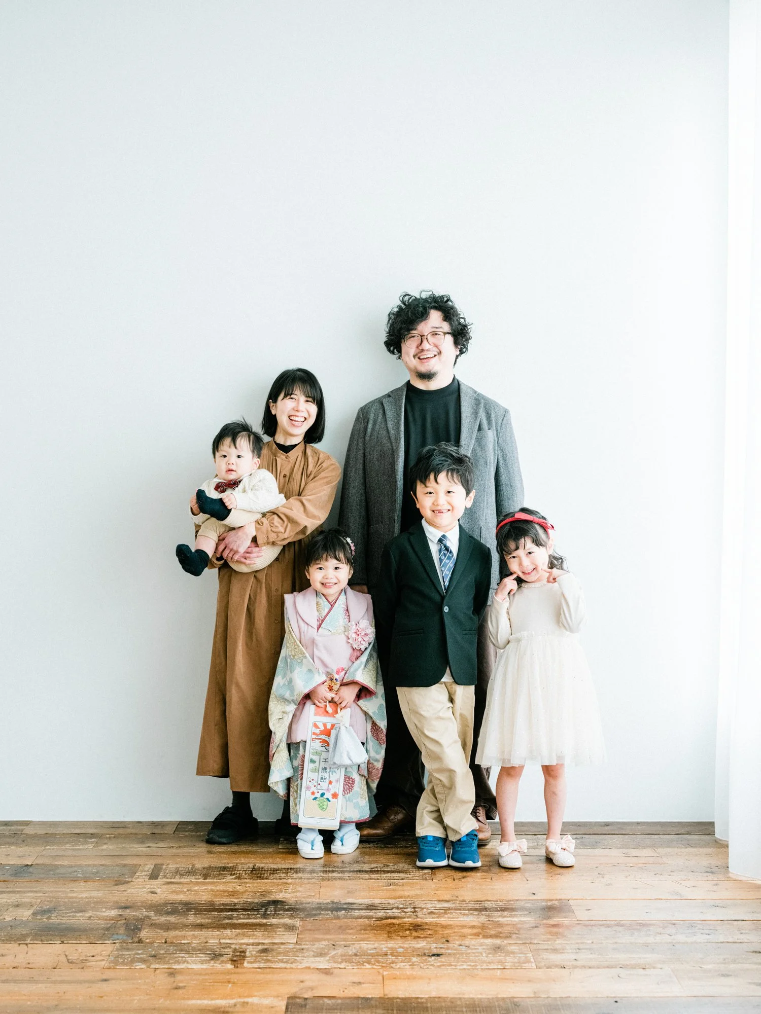 家族の写真。大人3人と子供4人が笑顔で並んでいる。背景は白い壁。