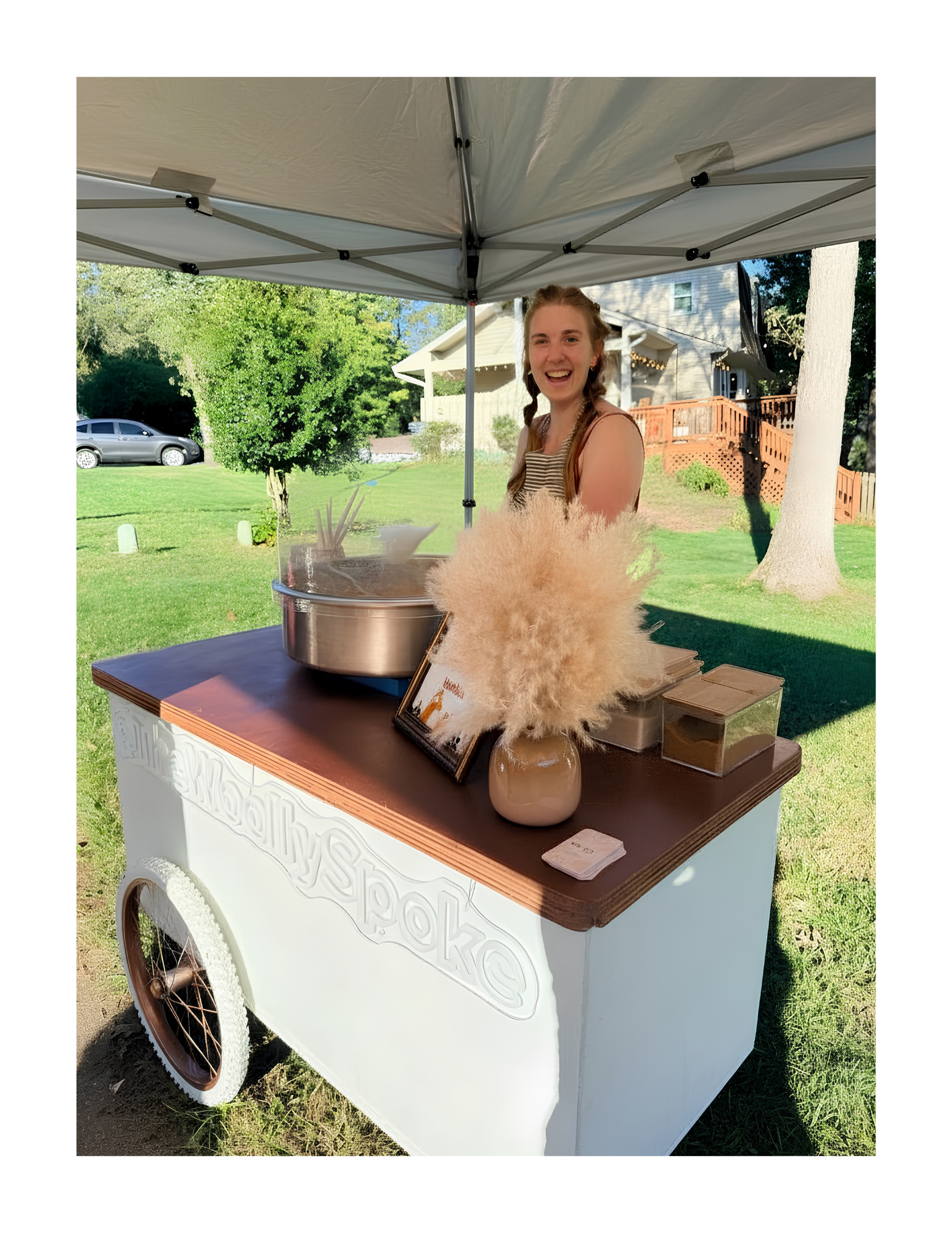A young woman with braided hair smiling behind a cotton candy stand under a canopy outdoors.