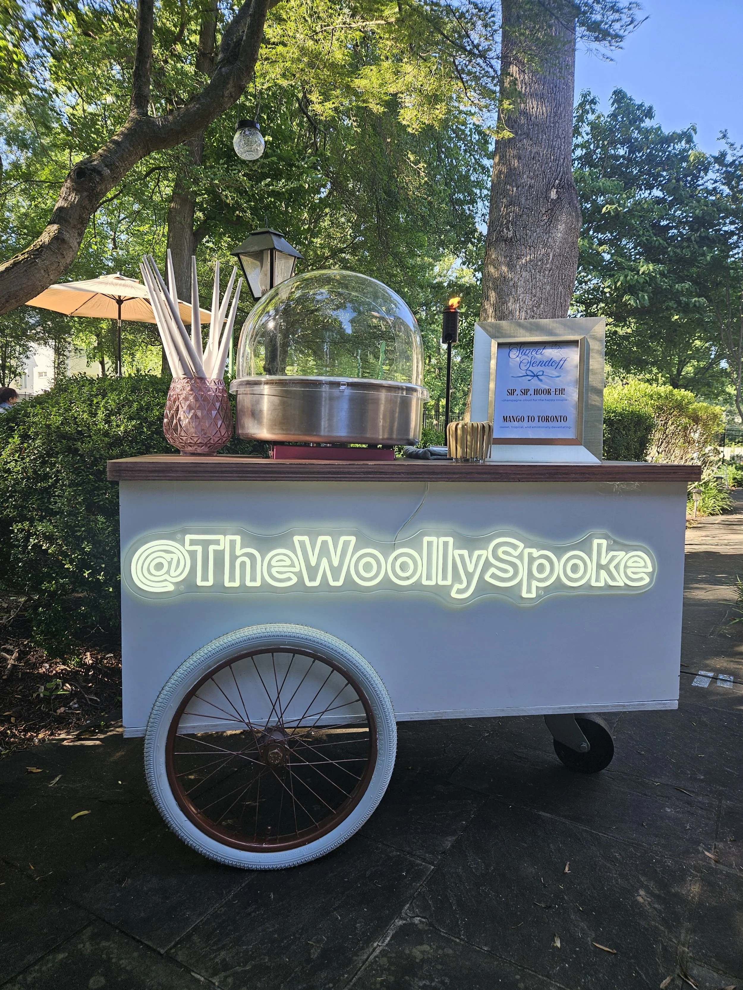 A white cotton candy cart with a large when and wooden countertop, decorated with a neon sign that reads ‘@TheWoollySpoke’ and a framed menu, set outdoors under trees on the club lawn.