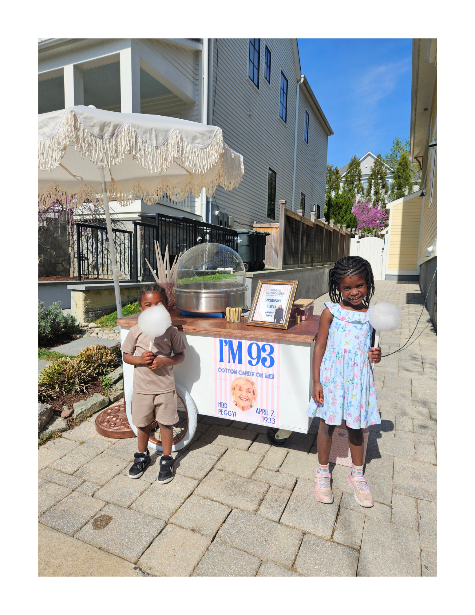 Two children standing outside next to a cotton candy cart under an umbrella during a sunny day in a residential area, with houses, trees, and a blue sky in the background. They are enjoying dye-free, organic cotton candy cones.
