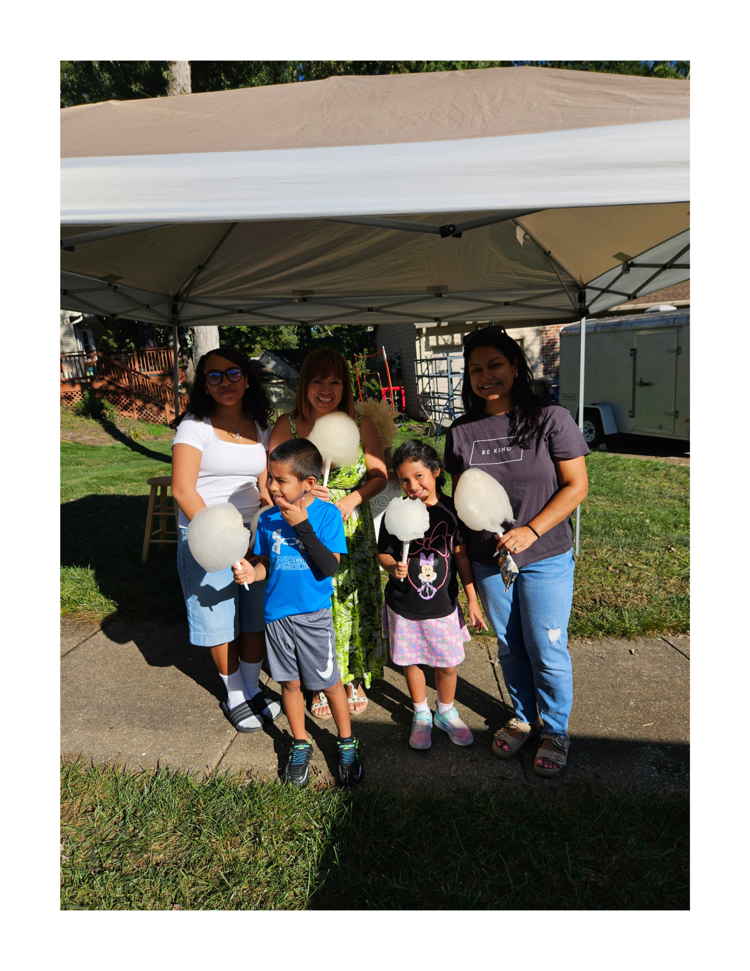 Group of five people, including children and adults, smiling and holding cotton candy at an outdoor block party gathering under a canopy, in a residential backyard during daytime.