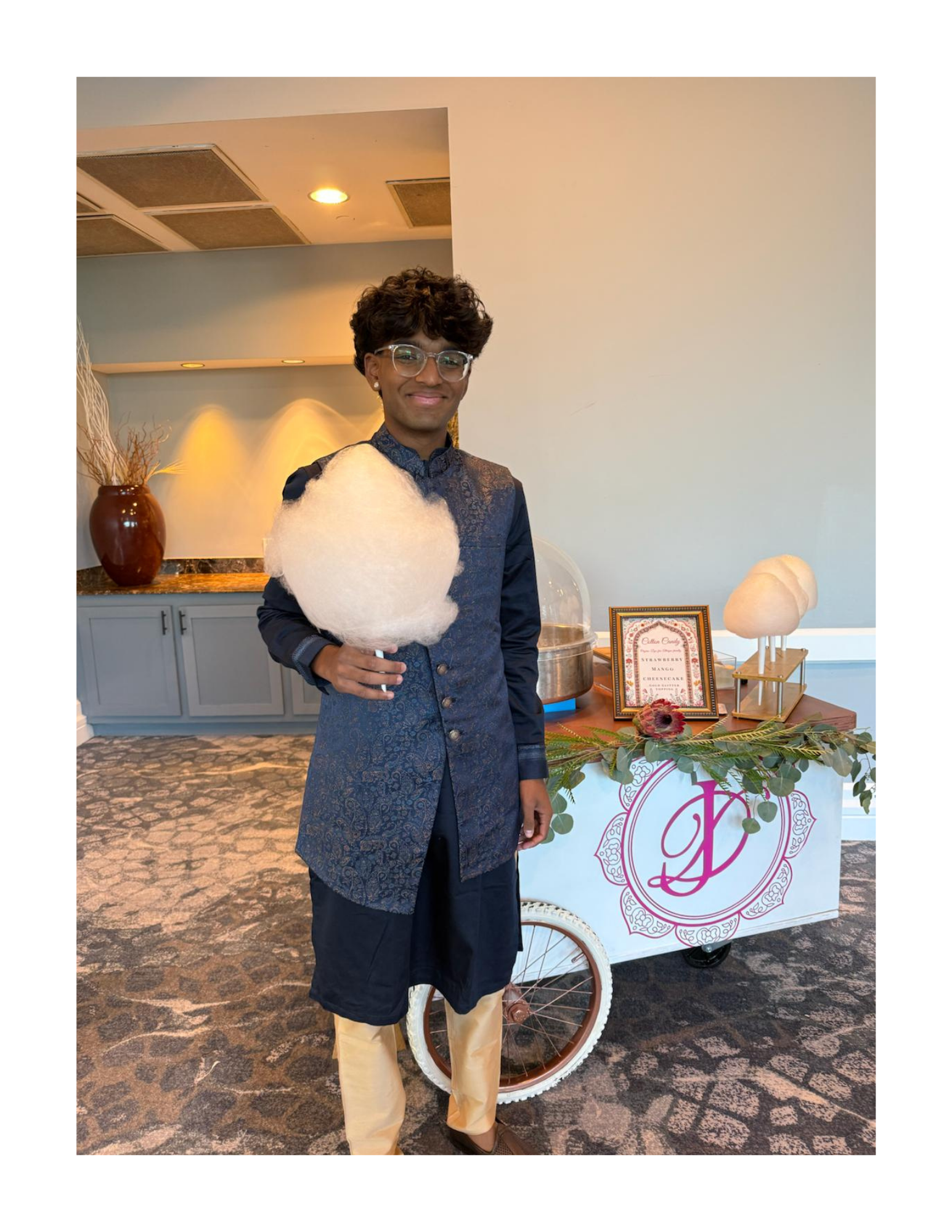 A young man dressed in traditional ethnic attire with glasses, holding a dye-free cotton candy cone with glitter, standing next to a decorated cart with a pink monogram and floral decorations, inside luxury banquet hall in Northern Virginia.