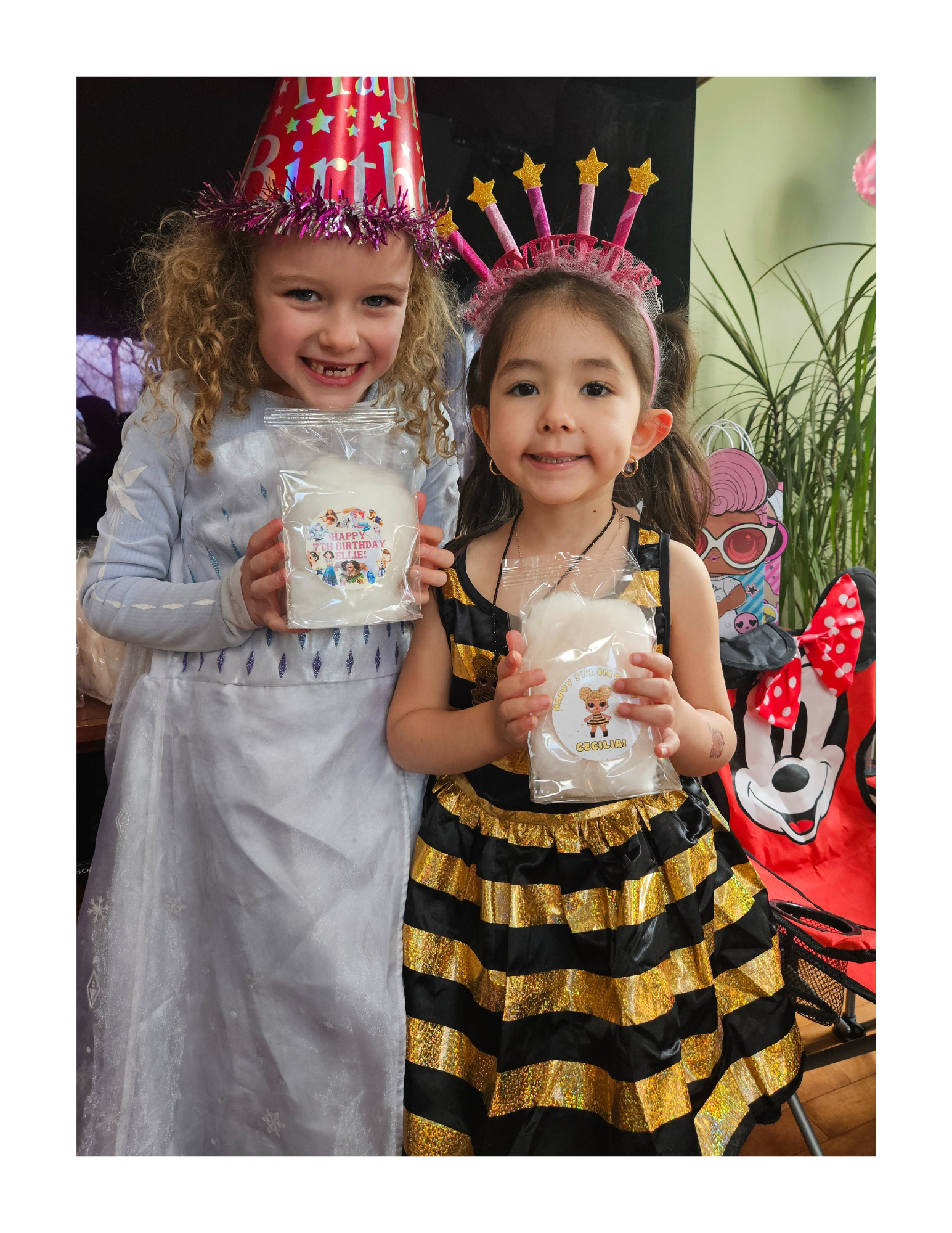 Two young girls at a birthday party wearing festive costumes and holding goodie bags. One girl is dressed as a princess in a white gown with snowflake designs and a pink birthday hat, while the other girl is in a black and gold dress with a pink headband with stars. They are smiling and celebrating indoors decorated with party items.