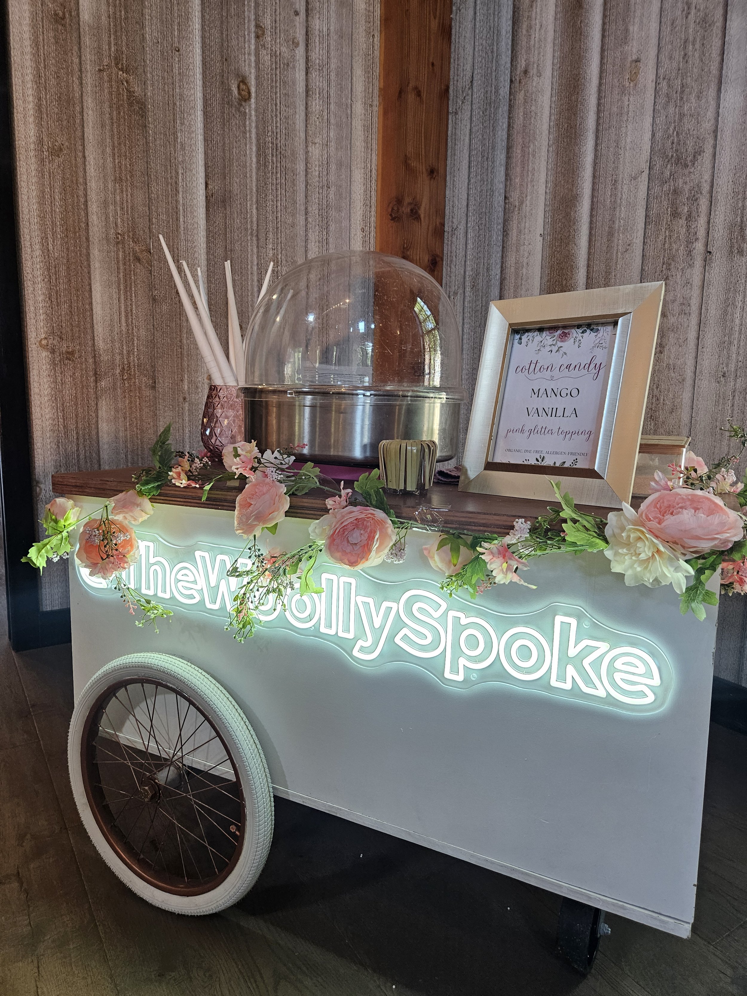 A cotton candy cart decorated with pink roses and greenery, with a neon sign that says 'the Holly Spoke.' The cart has white wheels with brown spokes, and a framed sign listing flavors including mango, vanilla, and pink glitter topping.