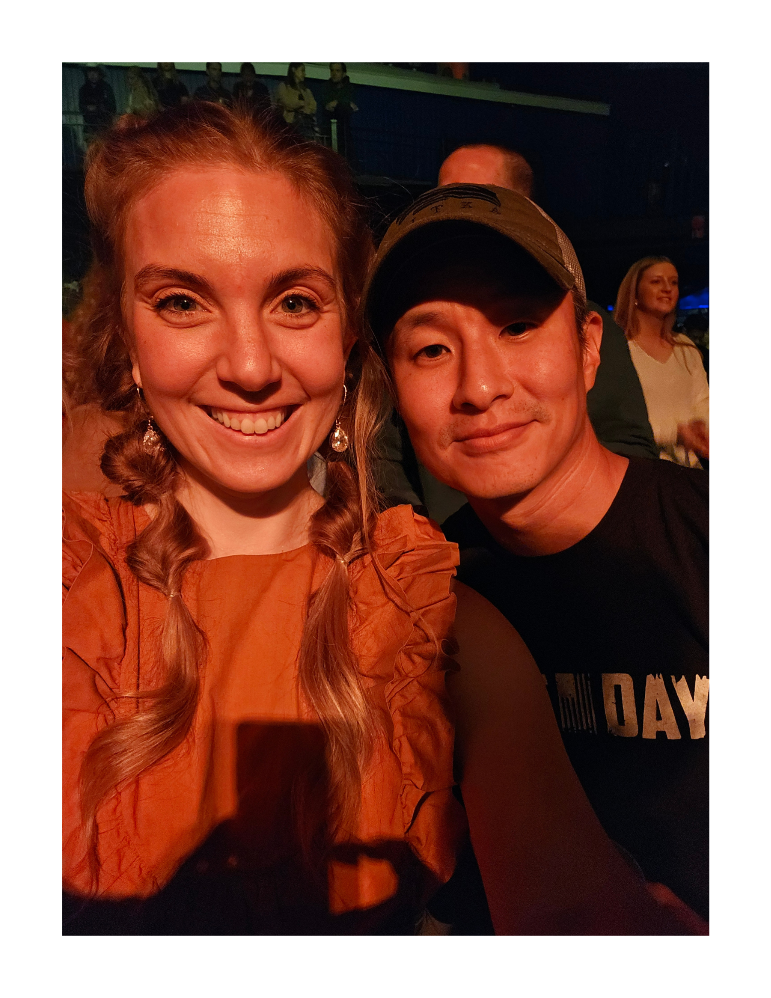 A smiling woman with long, wavy, reddish-brown hair and earrings, and a man wearing a baseball cap with a smile, posing for a selfie in a dimly lit indoor setting with other people in the background.