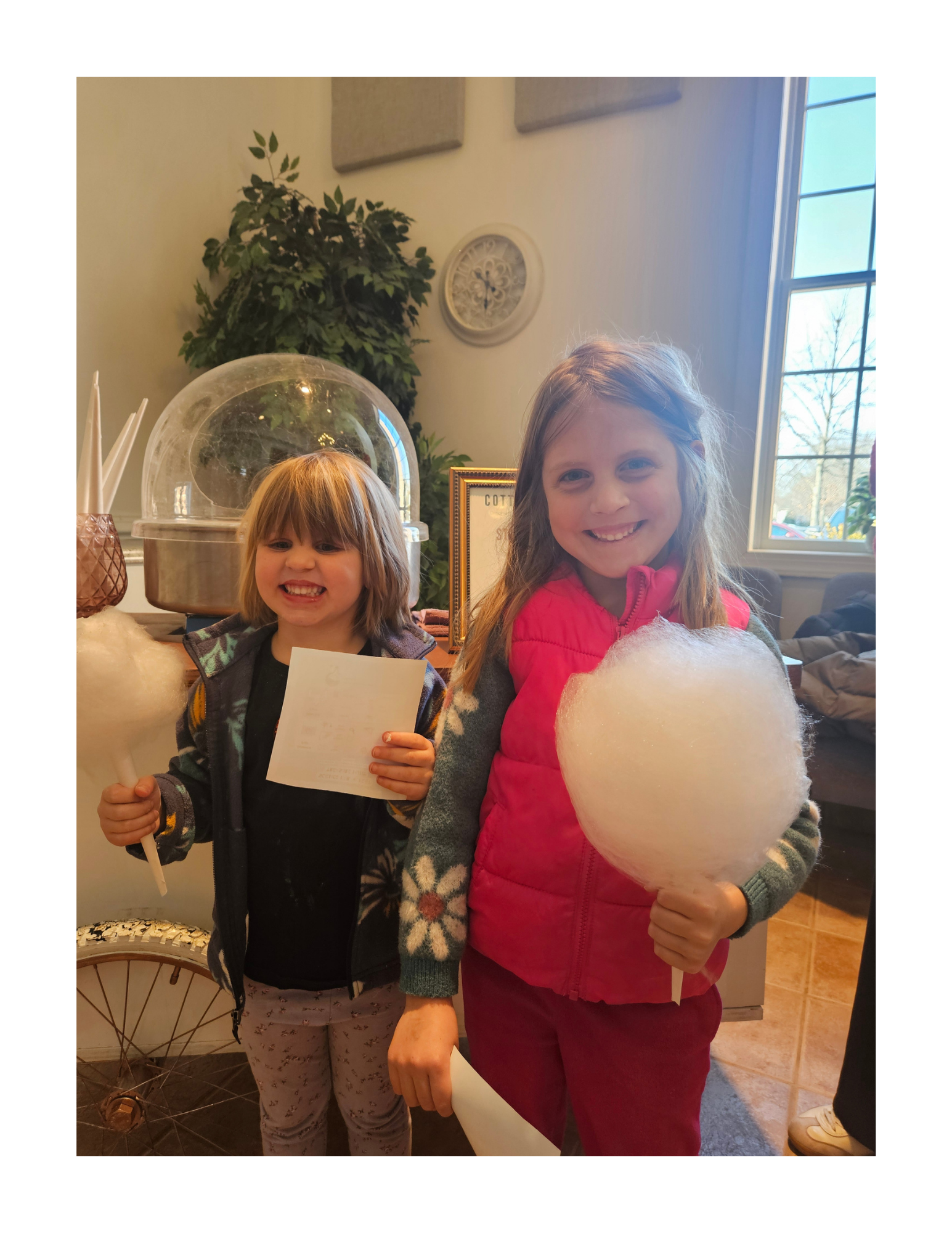 Two young girls smiling and holding cotton candy in a cozy indoor setting near a window, with decor and a bicycle wheel visible in the background.
