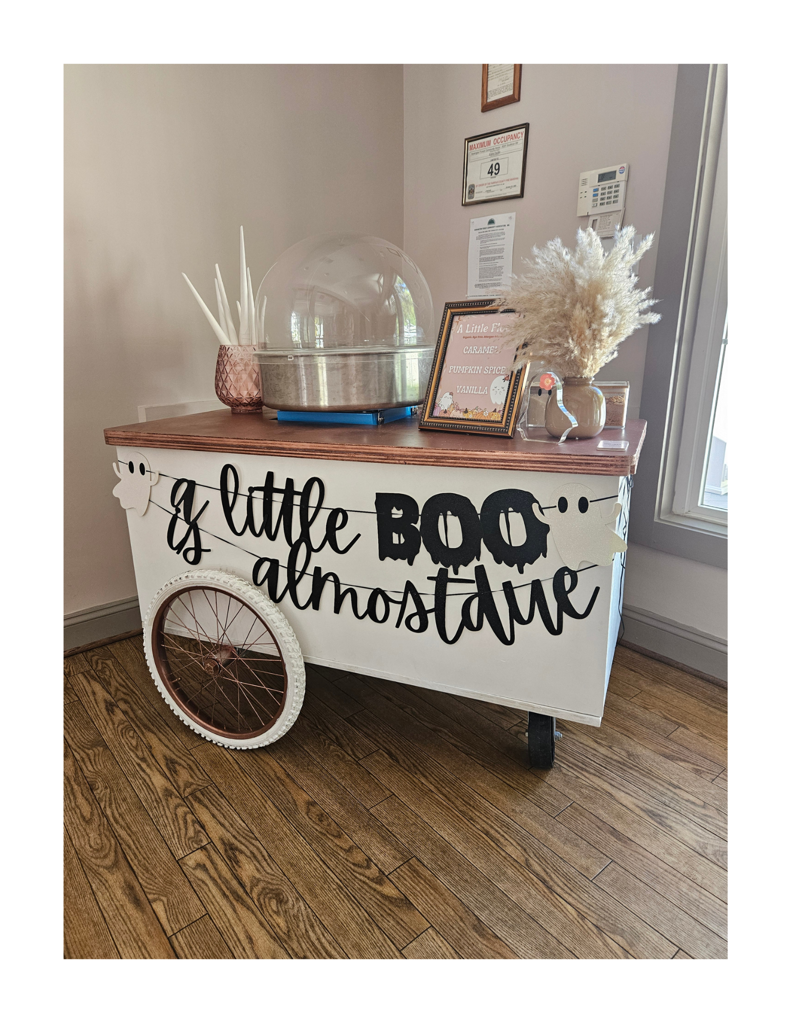 A custom cotton candy cart with black lettering reading 'A little BOO is almost due.' It has a rose gold wooden top with a pink textured vase holding white cones, a framed sign with fall flavors, and a vase with white fluffy pampas grass.