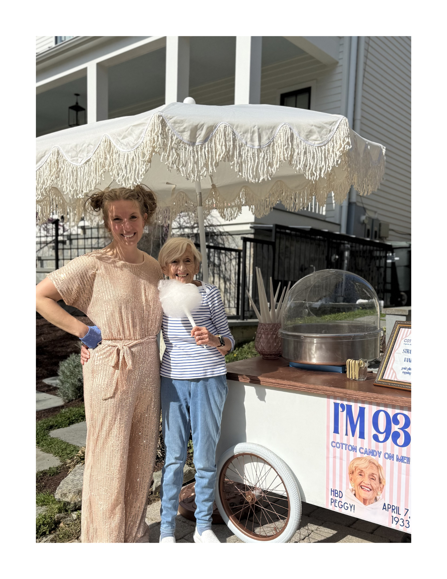 Two women standing on a sidewalk in front of a cotton candy stand, smiling at the camera. One woman has curly hair in a bun, wearing a beige dress, and the other has blond hair, wearing a striped shirt and jeans, holding cotton candy. The stand has a sign with a woman’s smiling face and the text "Cotton Candy on Me!" and birthday details for Peggy. There is a white parasol with fringe providing shade over them.