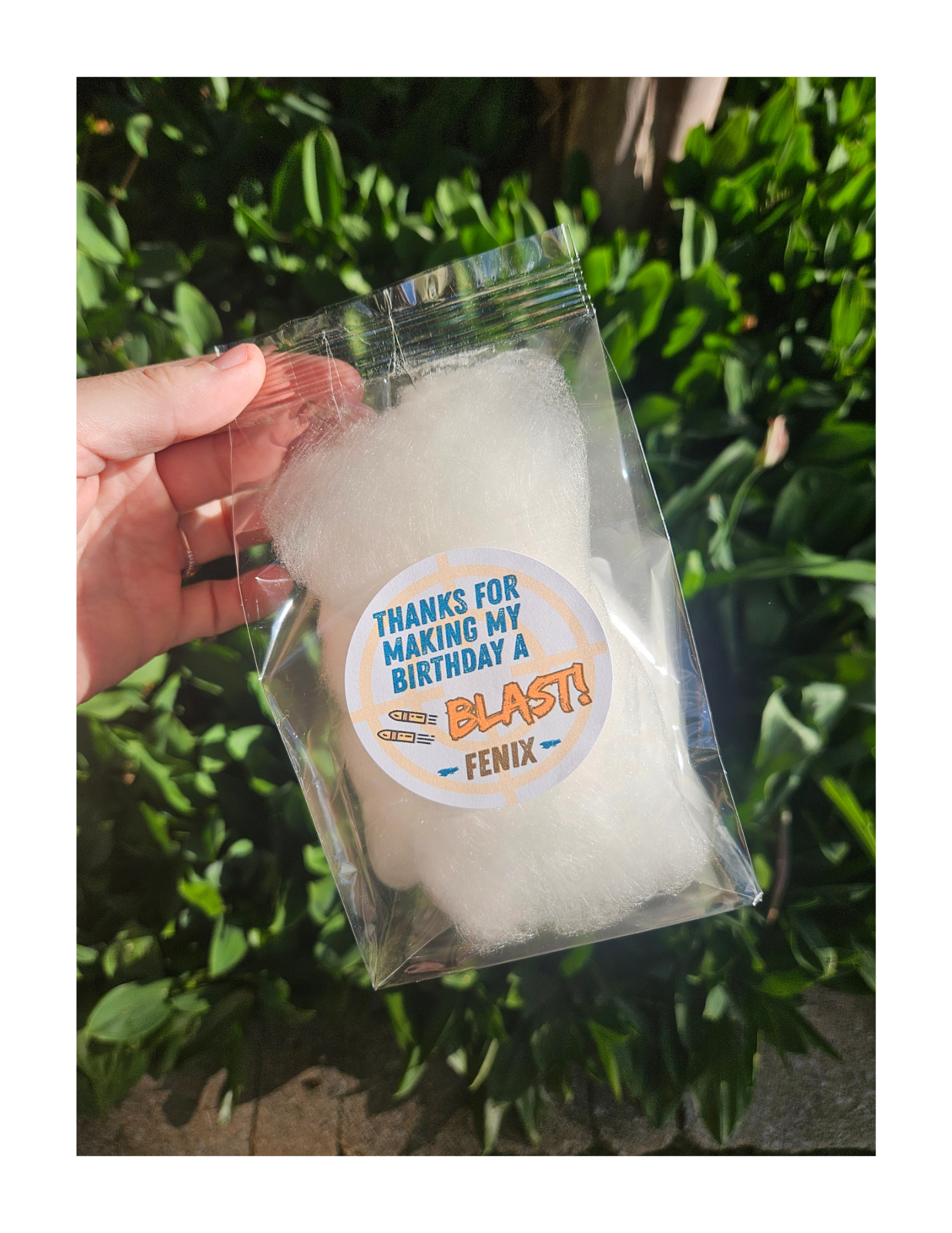 A hand holding a clear bag with dye-free cotton candy inside, outdoors with green leafy bushes in the background. The bag is a party favor for a boy's nerf gun themed birthday party.