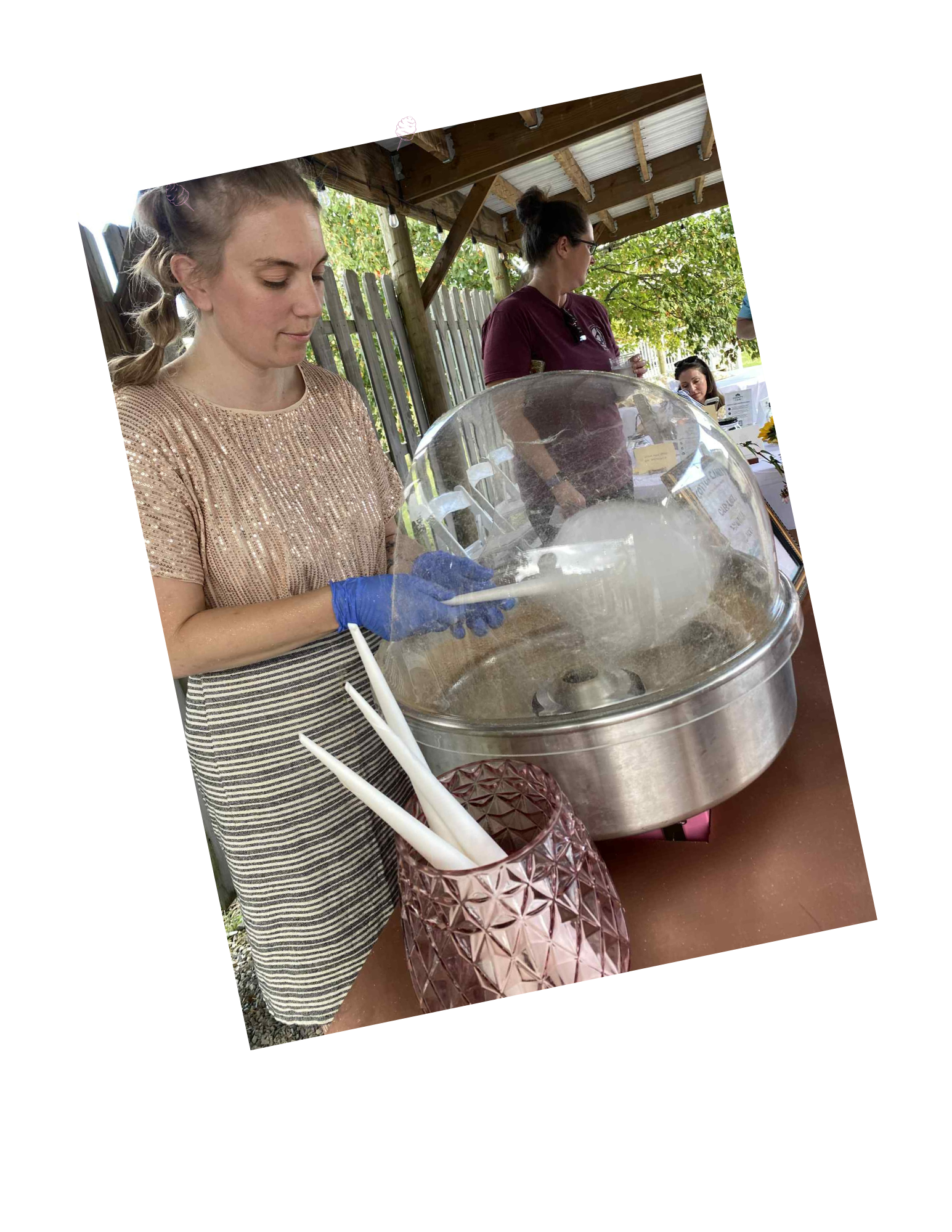 Women serving cotton candy at an outdoor event under a wooden pavilion with trees in the background.