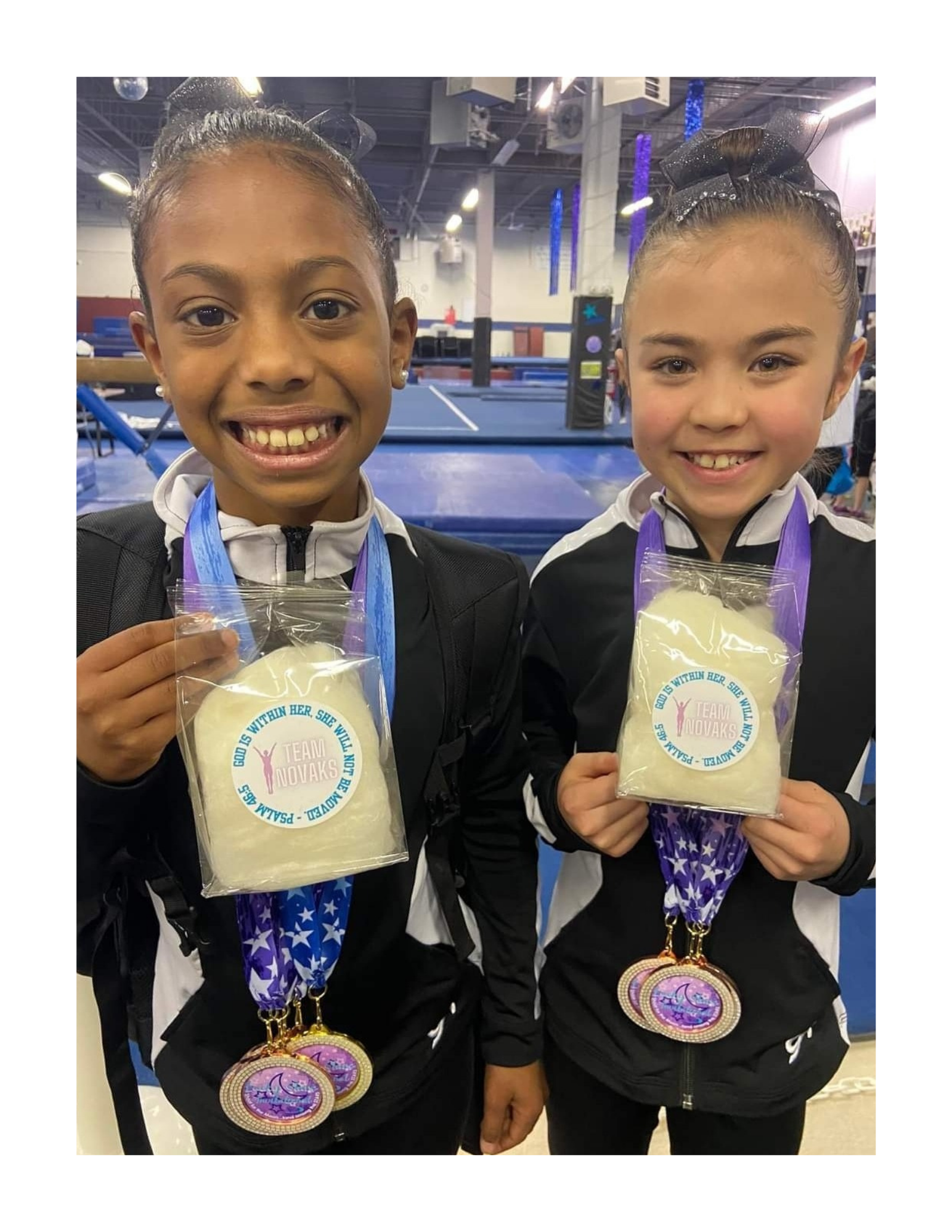 Two young girls in gymnastics uniforms smiling and holding awards, with medals hanging around their necks at a gymnasium.