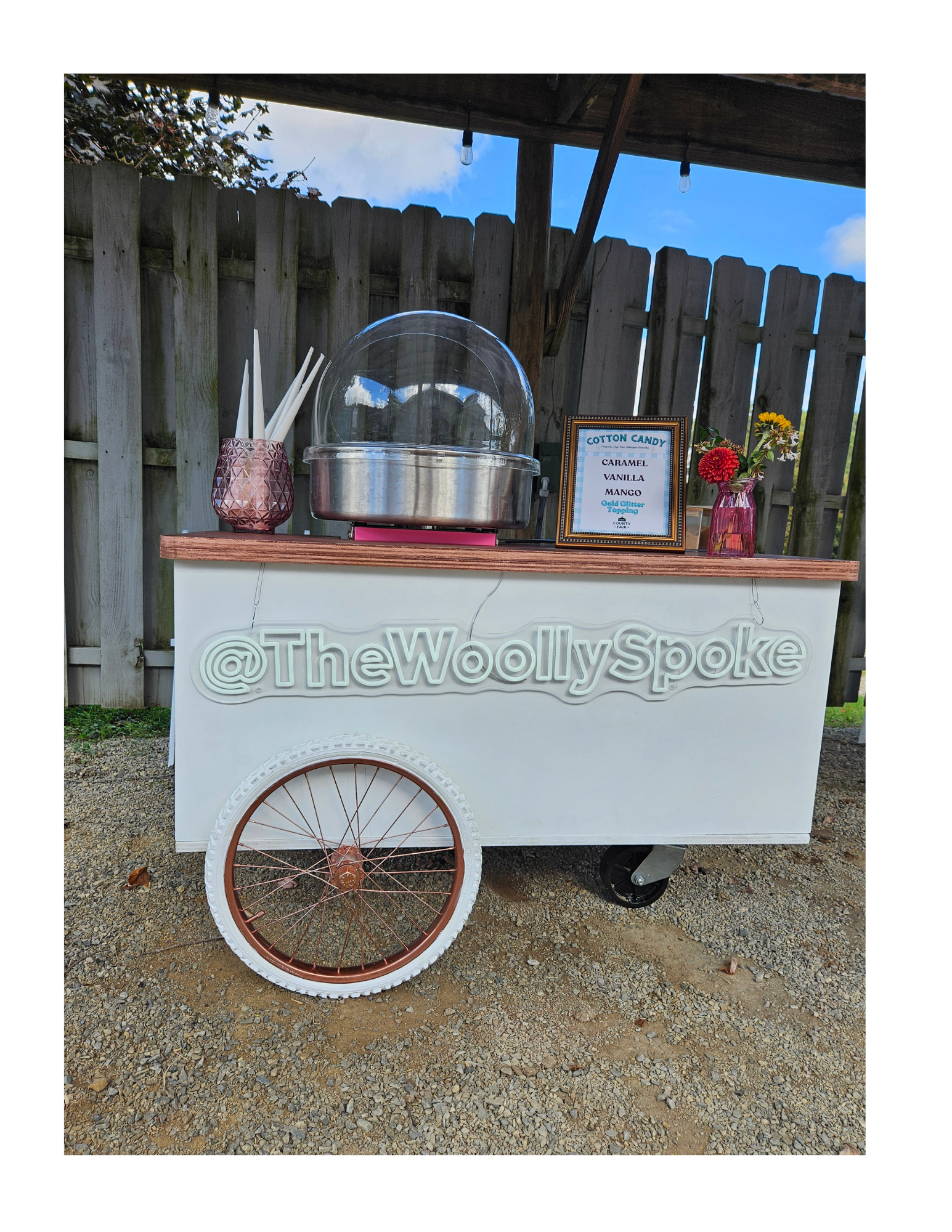 A white cotton candy cart with a sign displaying flavors including caramel, vanilla, and mango. The cart has a floral arrangement and a cup holding mini bamboo tongs. The cart has a large wheel with rose gold trim and is parked outdoors on a grave su
