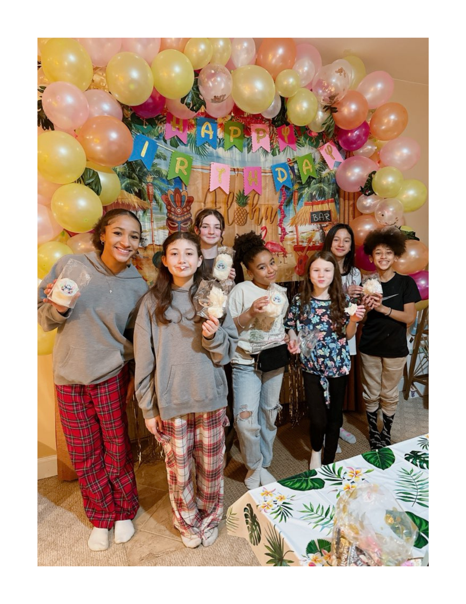 A group of girls at a birthday party, wearing pajamas and holding custom labeled, dye-free cotton candy bags , standing in front of a tropical themed backdrop decorated with balloons and a 'Happy Birthday' banner.