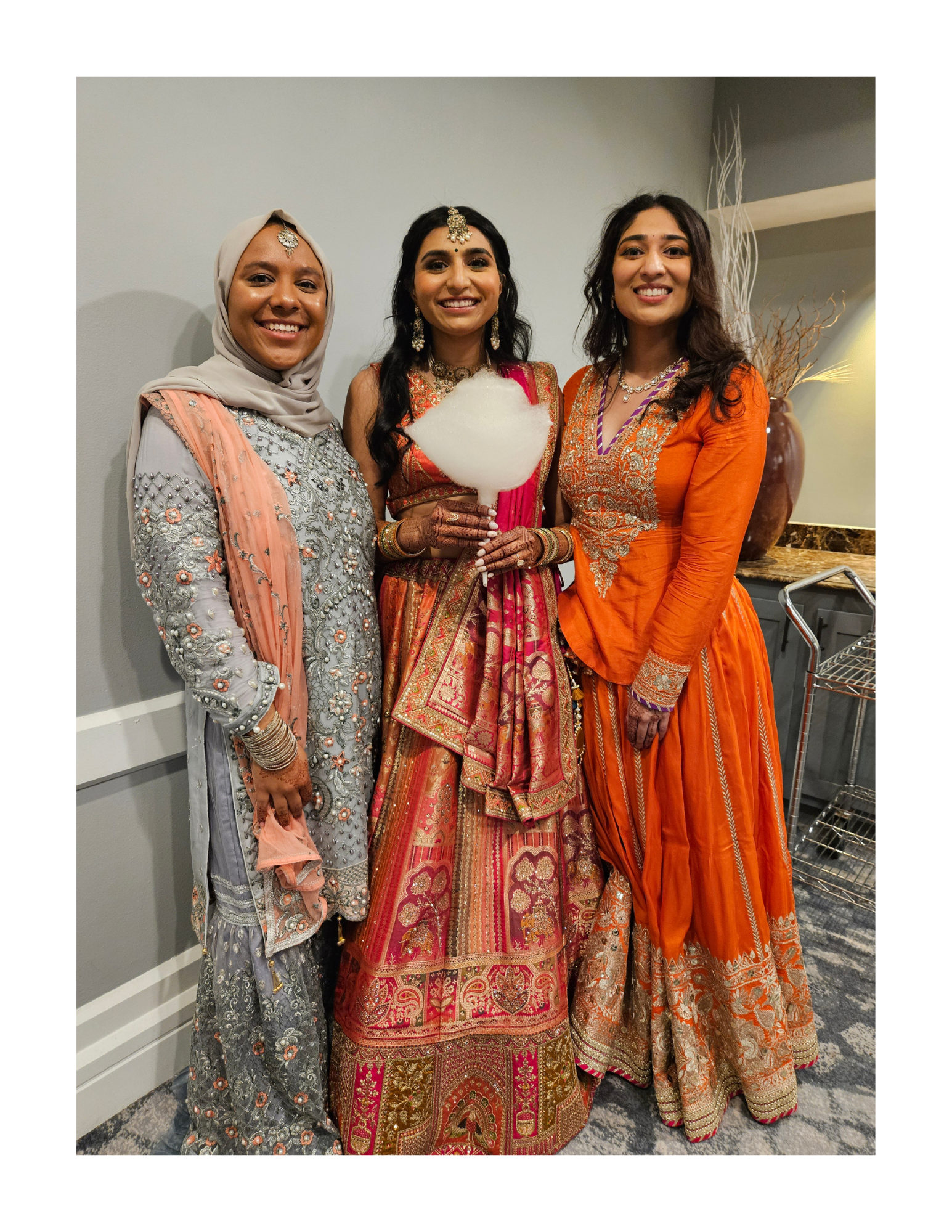 Three women dressed in traditional colorful Indian attire, smiling and standing together indoors eating dye-free, organic cotton candy cones.