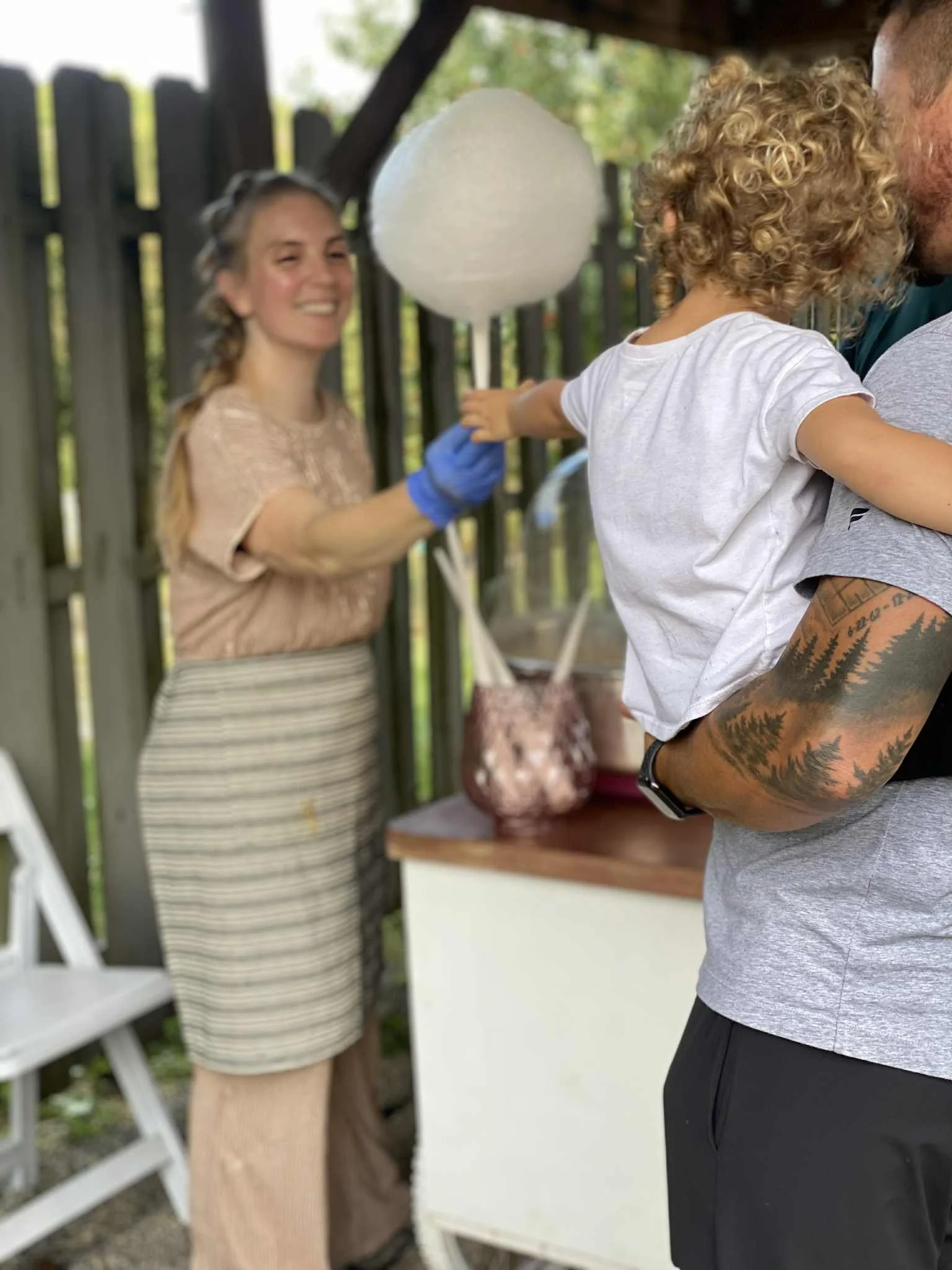 A man with tattoos on his arm holding a young child with curly blonde hair, at an outdoor gathering with a woman smiling in the background handing them a dye-free cotton candy cone with edible glitter.