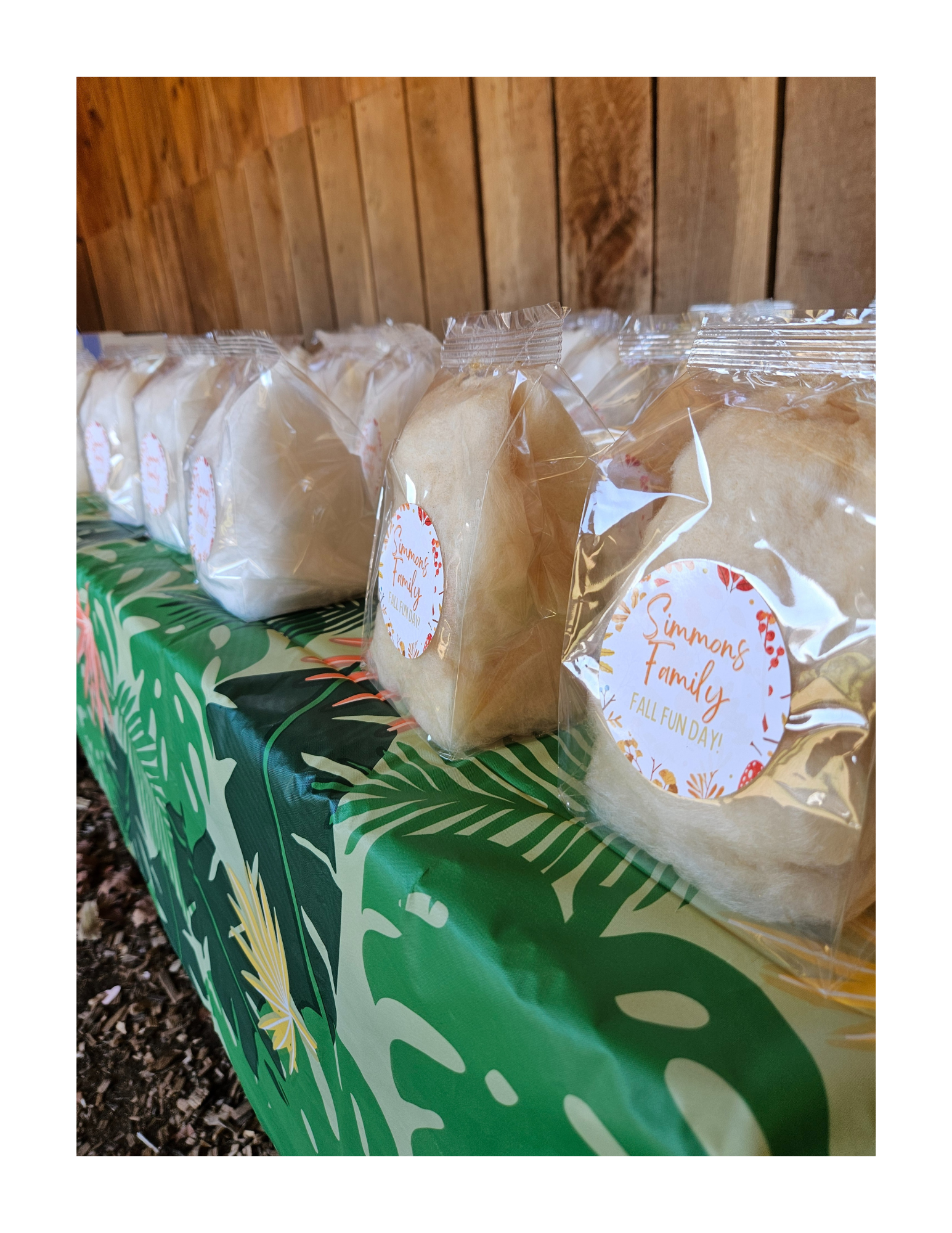 Multiple clear gusseted bags of cotton candy with a 'family reunion' sticker on a table with a tropical patterned tablecloth, against a wood-paneled wall.
