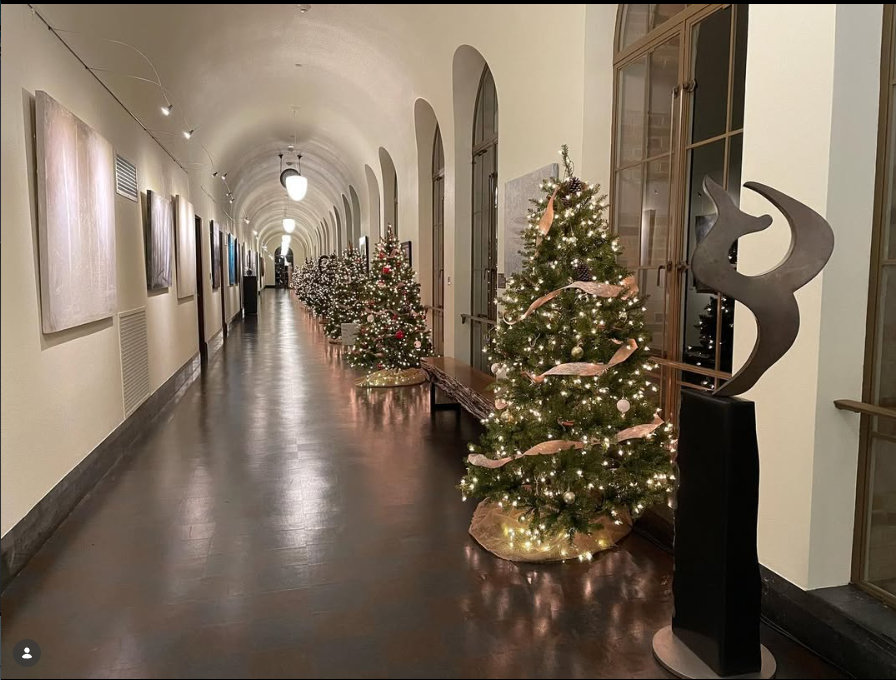 Decorated Christmas trees with lights and ribbons along a hallway with arched windows and artwork on the wall.