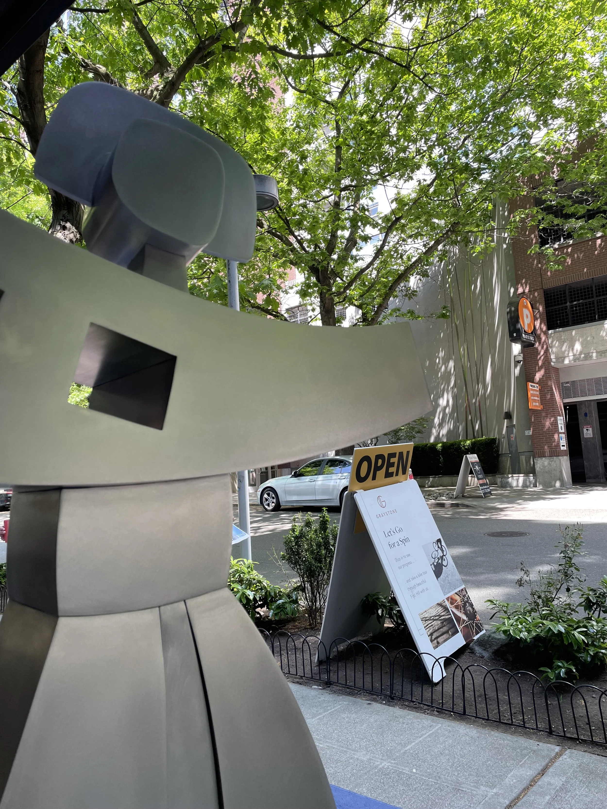 Street scene showing a modern abstract sculpture in the foreground, a green leafy tree, a parking sign, and a building with signs that read "OPEN" and other informational boards in the background.