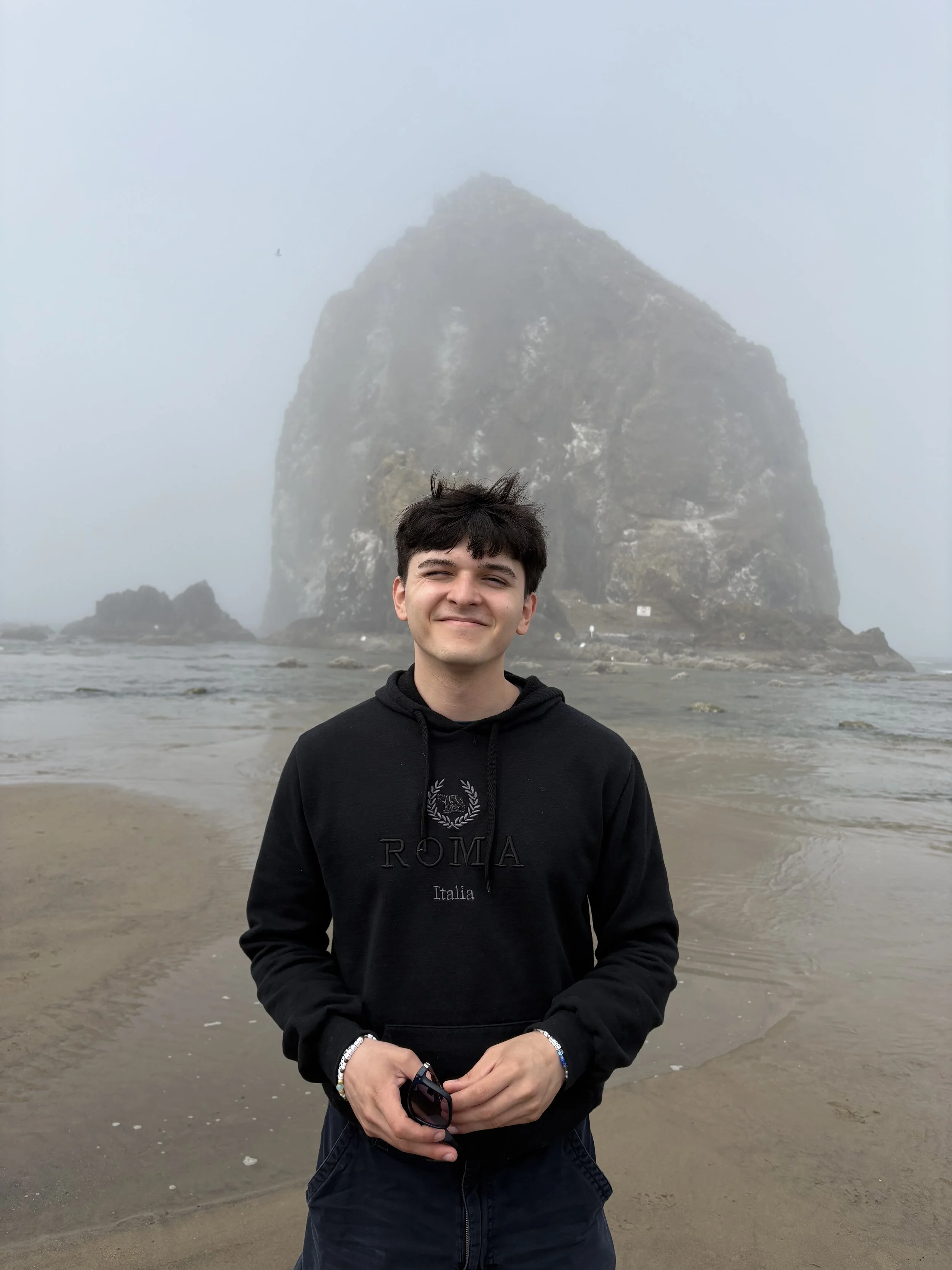 A young man with dark hair smiling and standing on a beach, holding sunglasses, with a large rock formation and misty sky in the background.