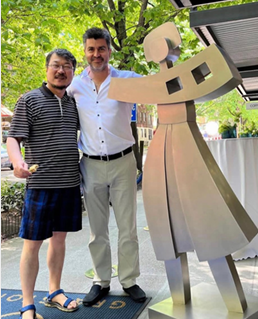 Two men standing outdoors next to a large metallic sculpture of a woman. The man on the left wears glasses, a striped shirt, and shorts, while the man on the right wears a white shirt and light-colored pants. They are smiling, with greenery in the background.