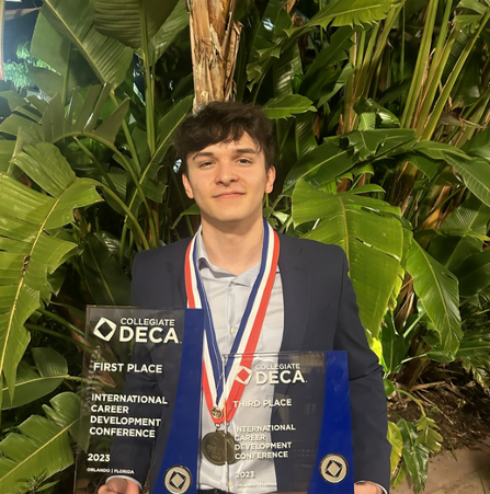 Young man in blazer with medals and awards, holding two trophies that read 'First Place' and 'Third Place' at the International Career Development Conference, with green tropical plants in the background.