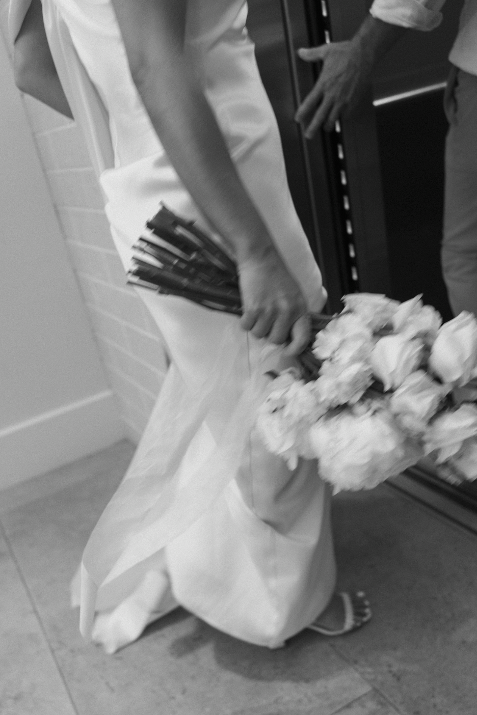 A person in a white gown holding a bouquet of white flowers, walking indoors near a brick wall, with another person's hand reaching out.