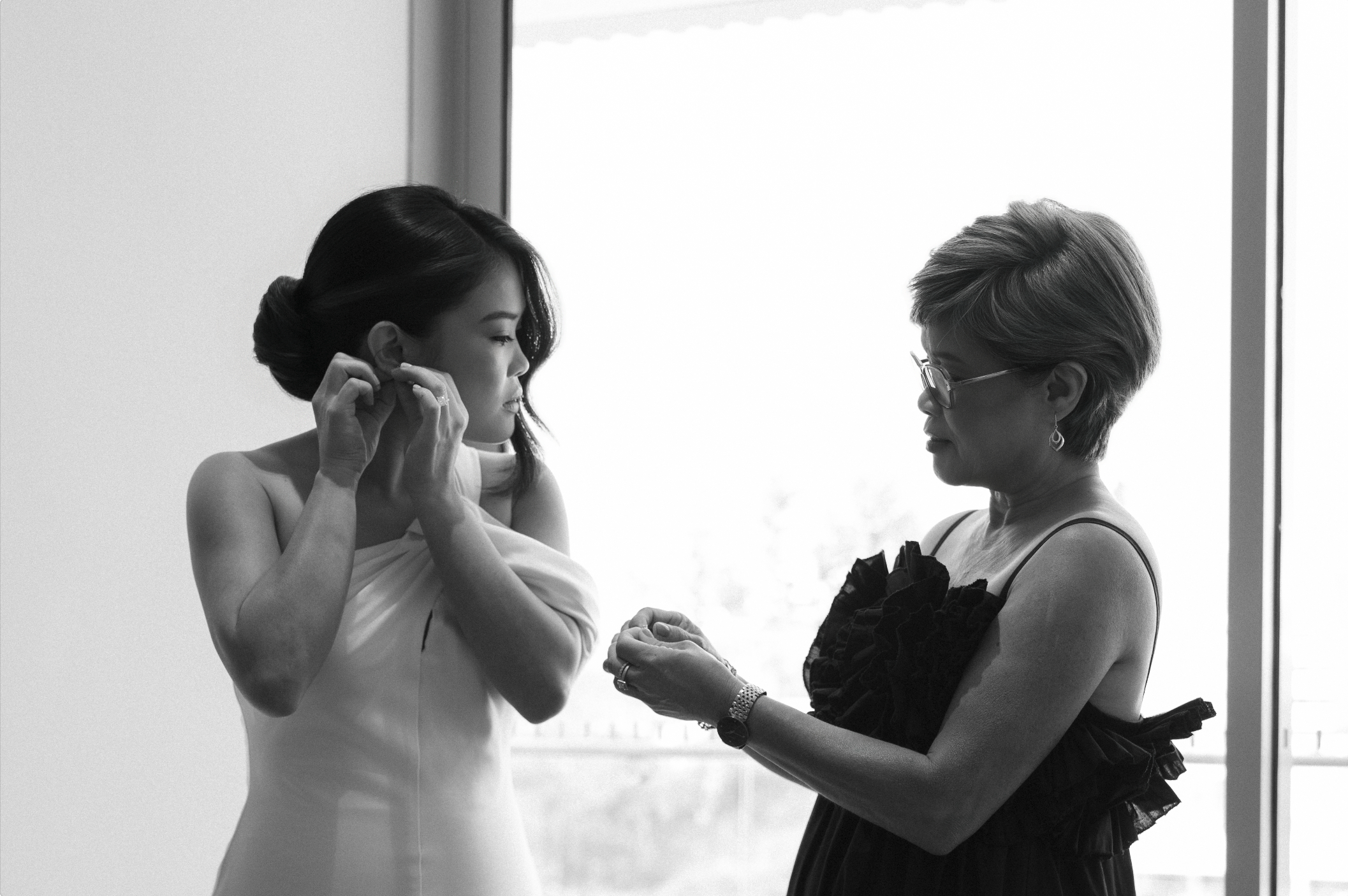 A woman with short hair and glasses is presenting a ring to a bride, who is adjusting her earring, during a wedding ceremony in front of a large window.