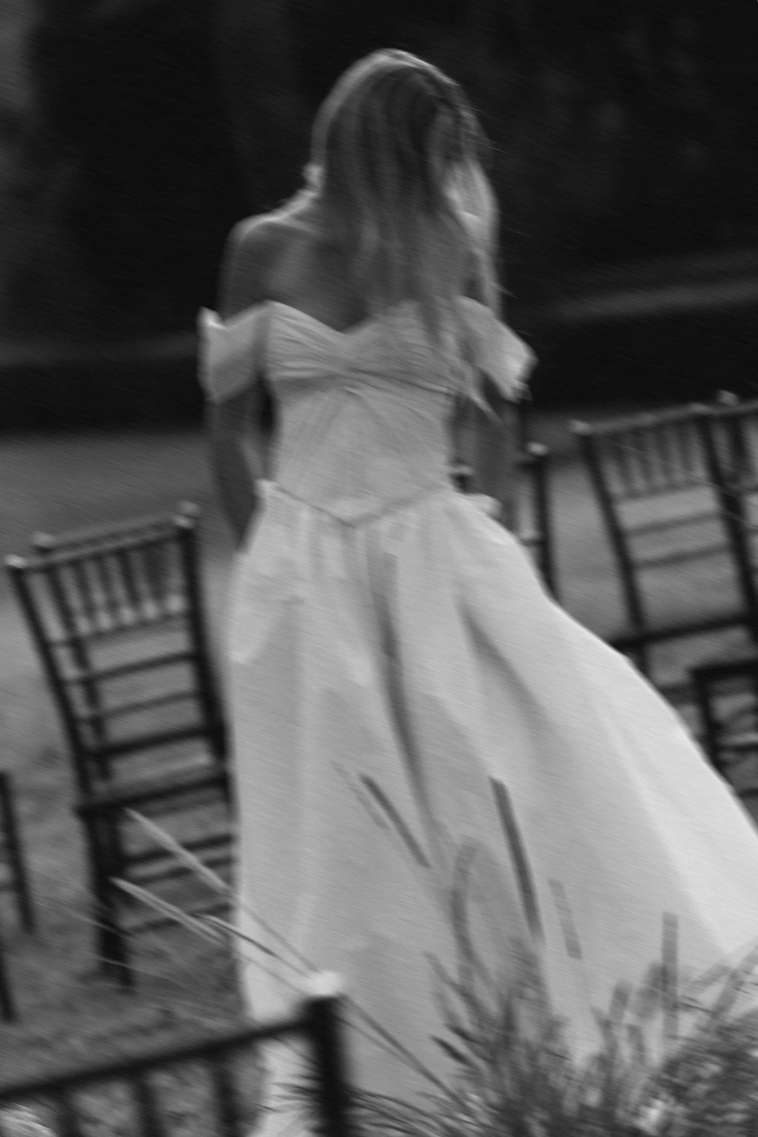 Blurred black-and-white photo of a woman in a strapless white dress, walking outdoors between outdoor chairs.