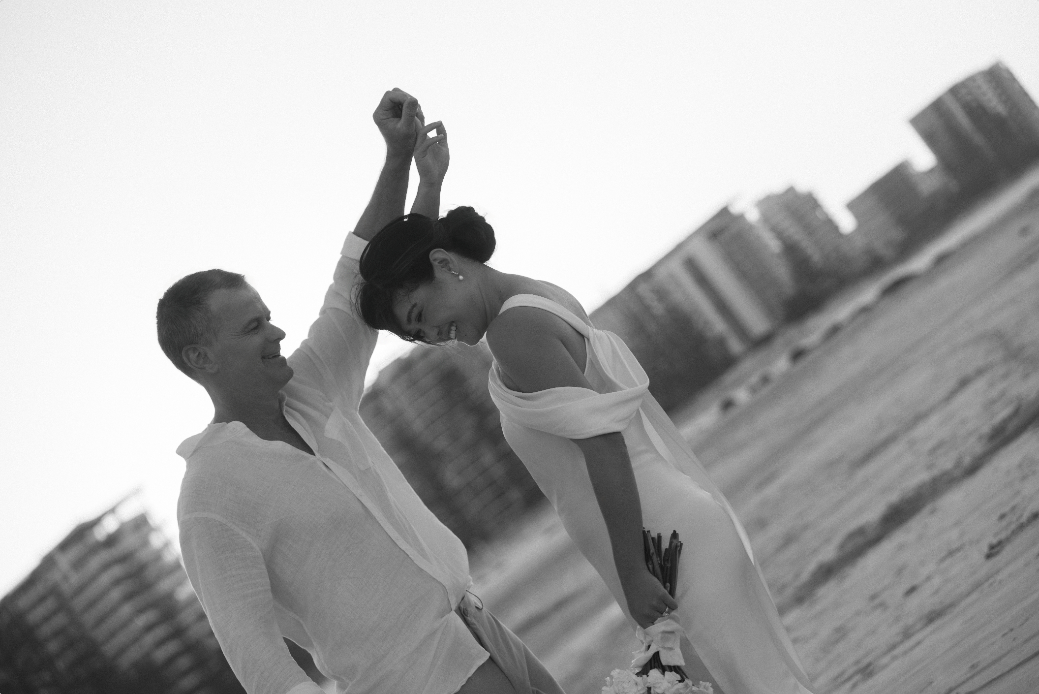 A black and white photo of a couple holding hands, smiling, with the city skyline in the background.