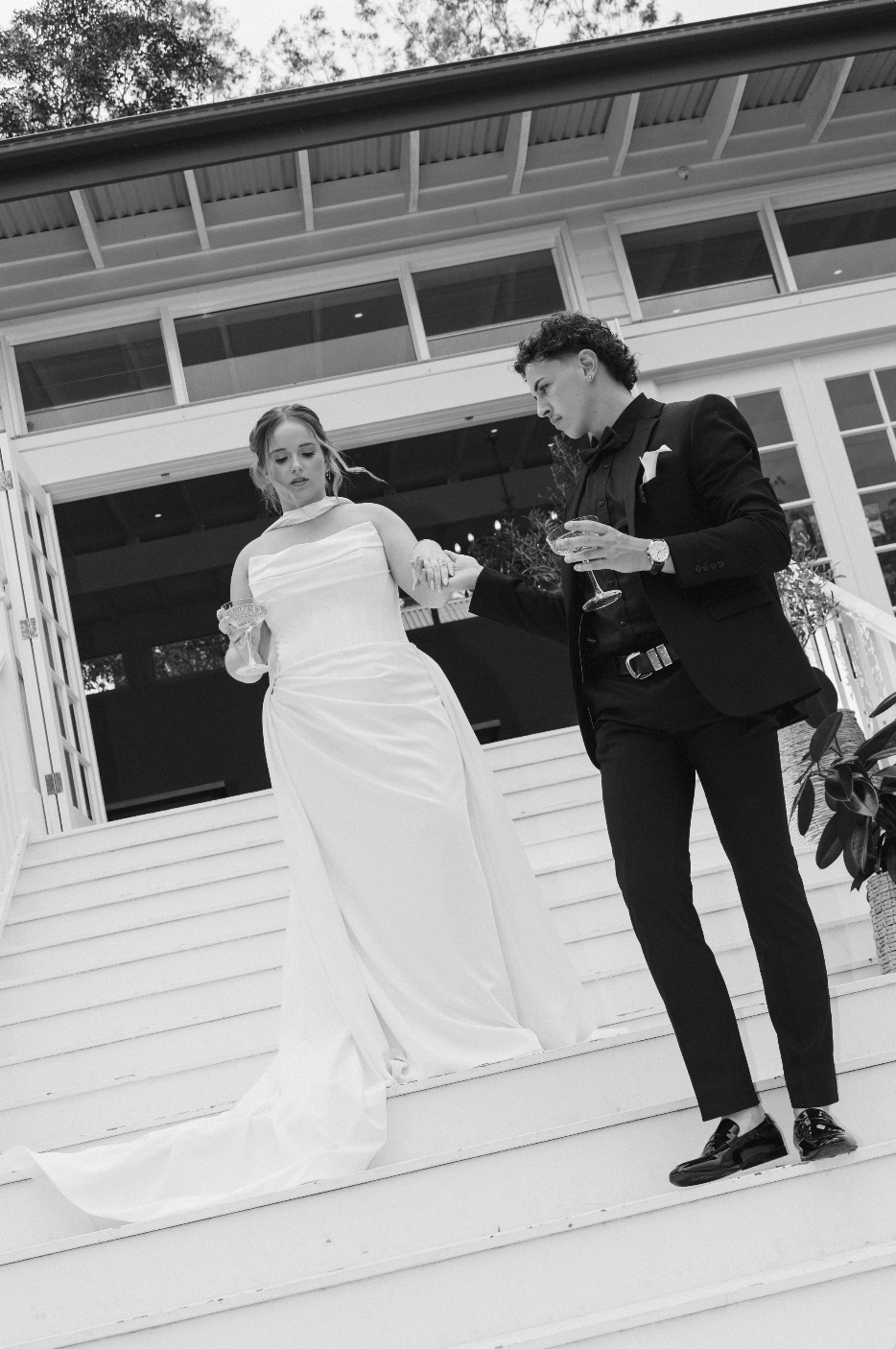 A black-and-white photo of a bride and groom standing on steps outside a house. The bride is wearing a long white gown, holding a glass, and looking down, while the groom is dressed in a black suit, holding the bride's hand, and looking at her.