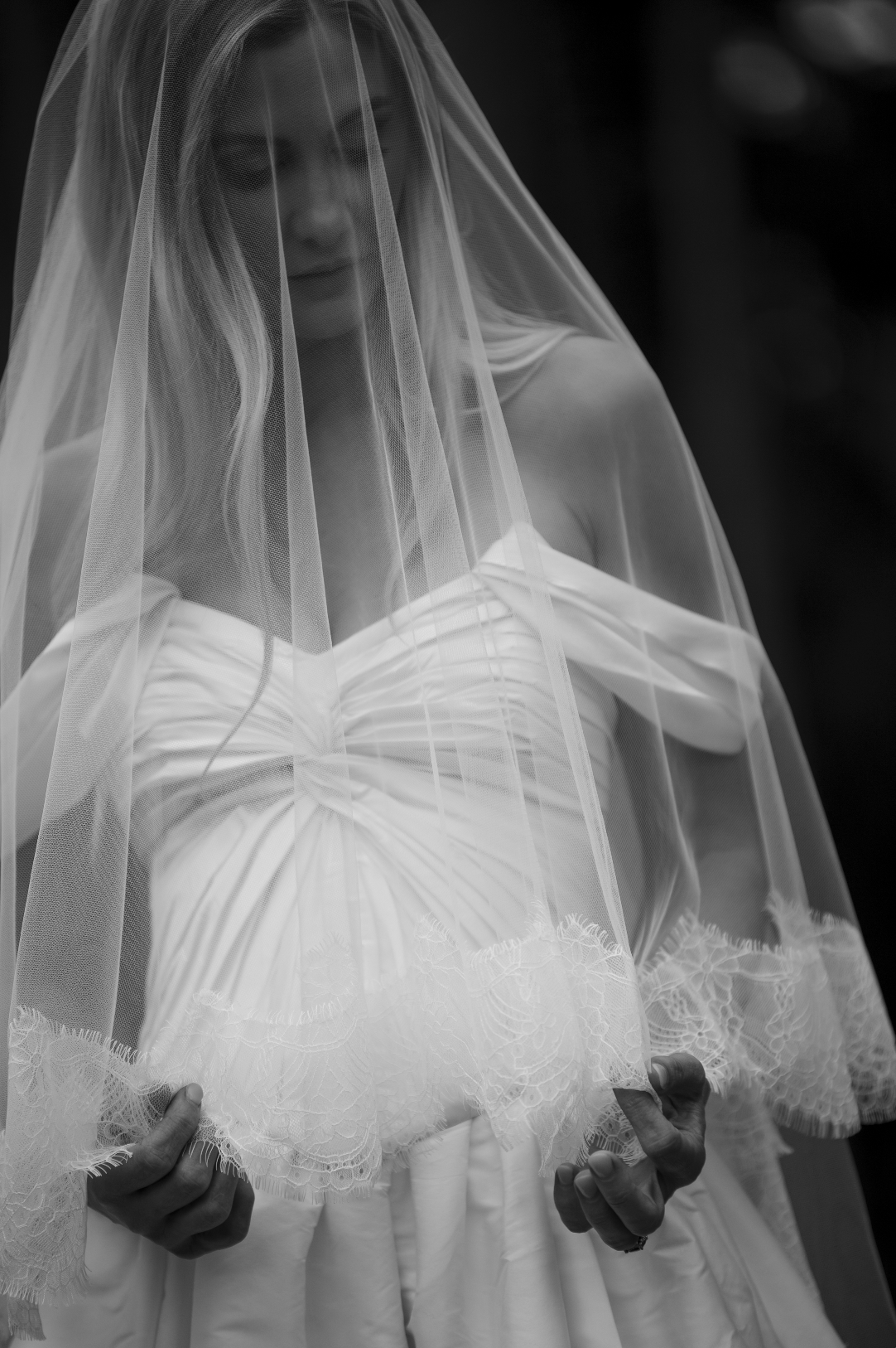 Black and white photo of a woman in a wedding dress and veil, partly covering her face, with her hands holding the dress.