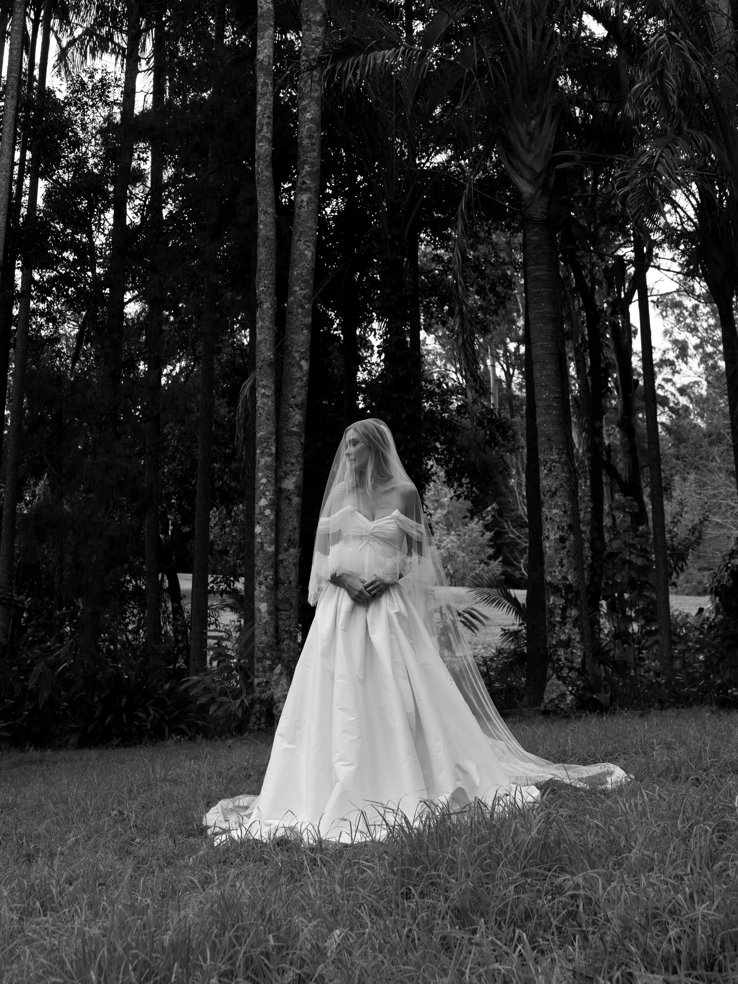 A woman in a wedding dress and veil standing outdoors among trees and grass.