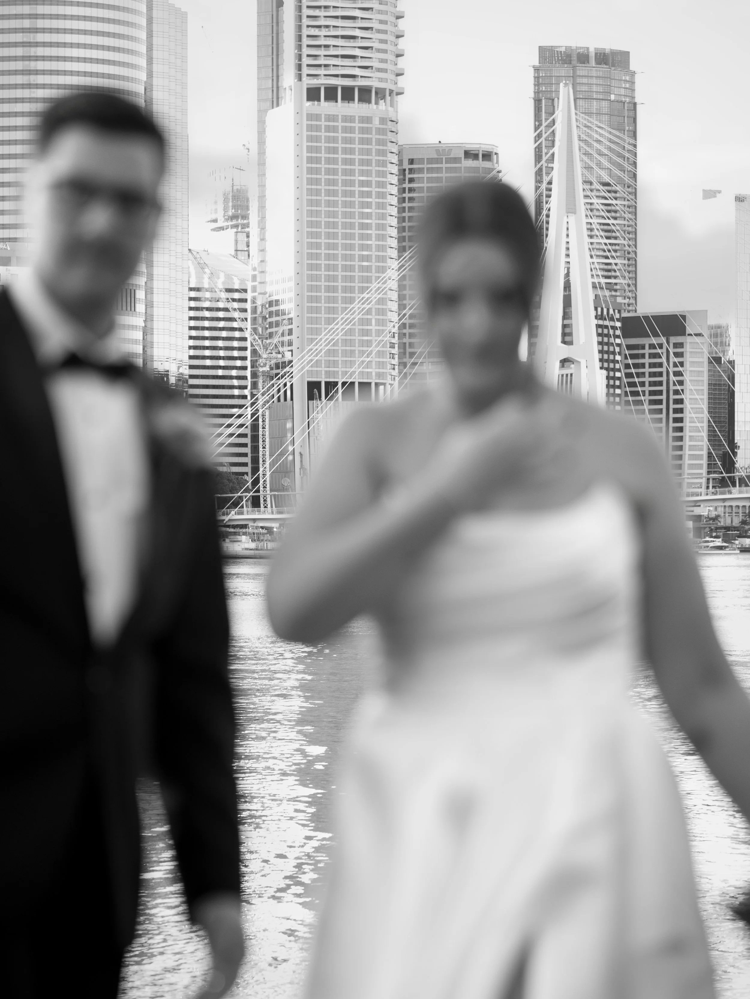 Blurry image of a man in a tuxedo and a woman in a white dress with city skyscrapers and a bridge in the background.