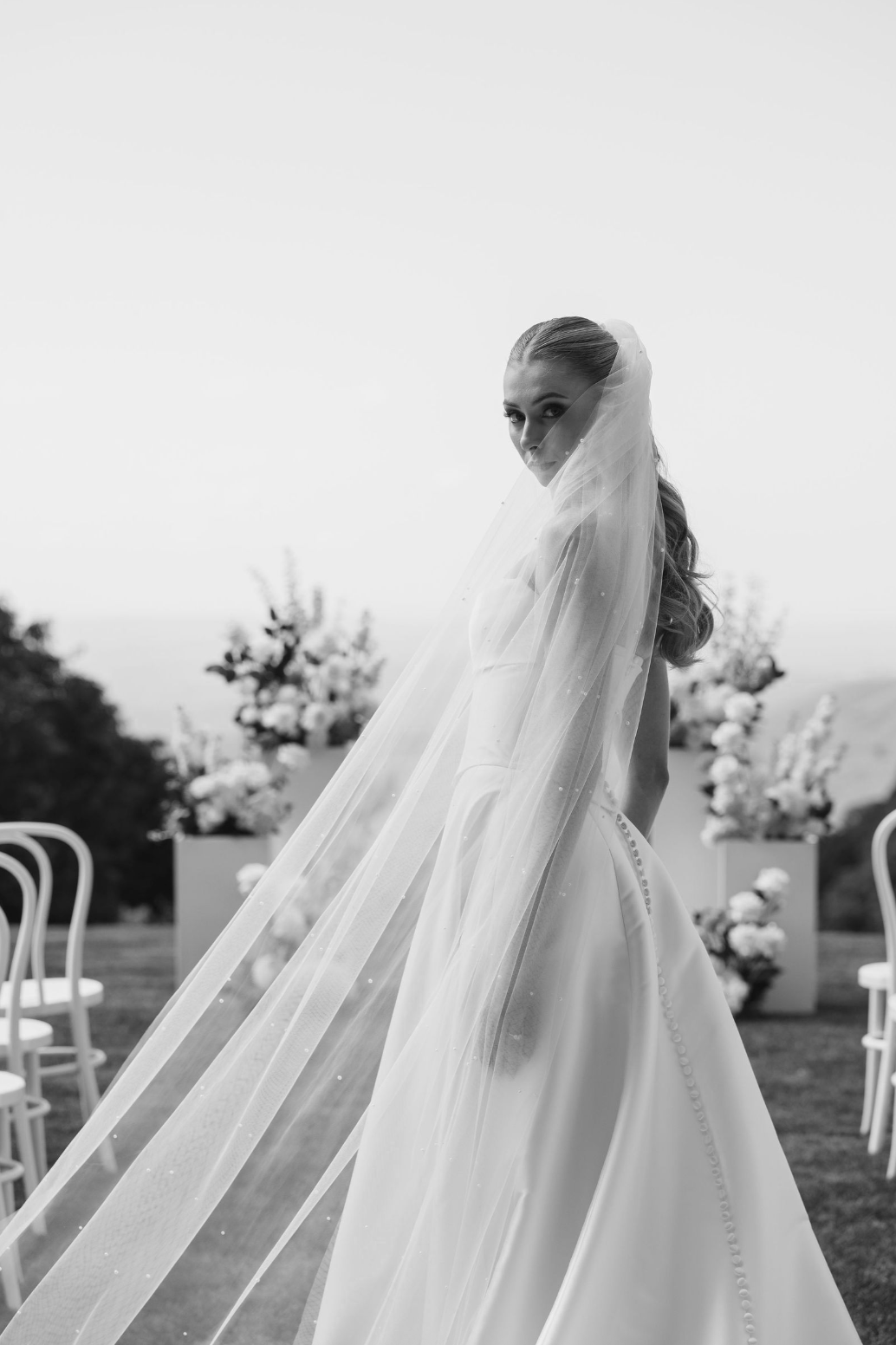 Black and white photo of a bride with a veil, standing outdoors with chairs and floral arrangements in the background.