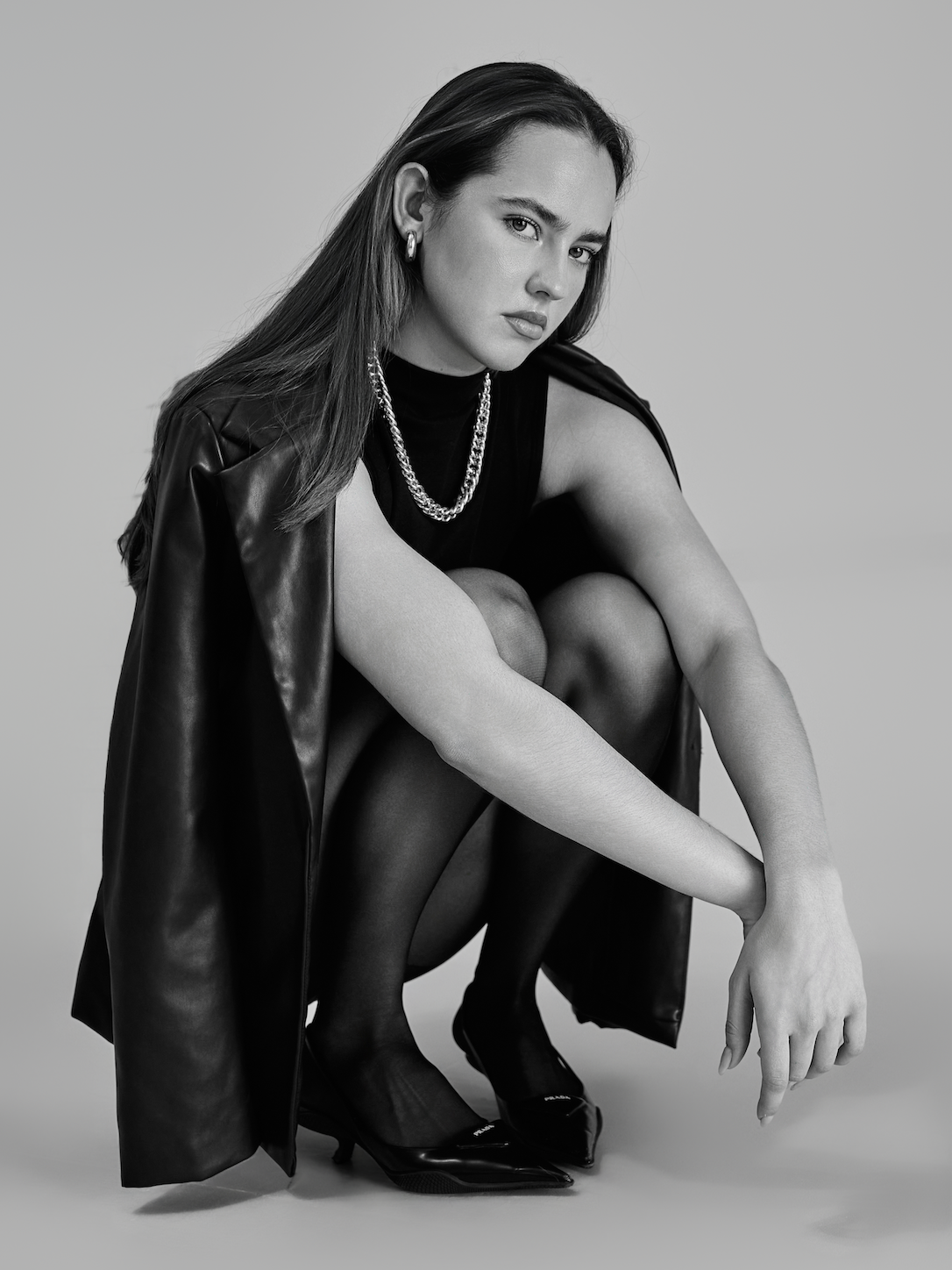 A black and white photo of a woman squatting, wearing a sleeveless black top, shiny black heels, a leather jacket draped over her shoulder, and jewelry including a chain necklace and hoop earrings.