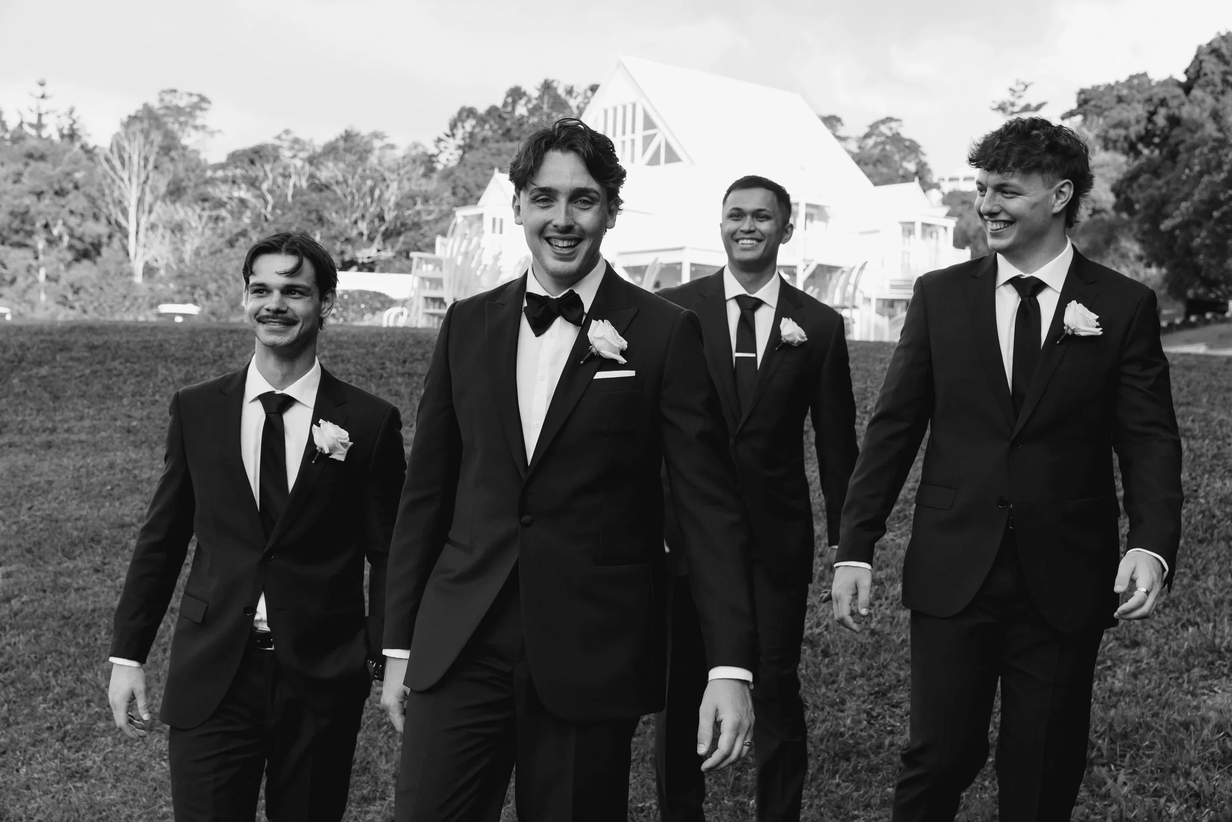 Four men in tuxedos walking outdoors, smiling, with a modern white house and trees in the background.
