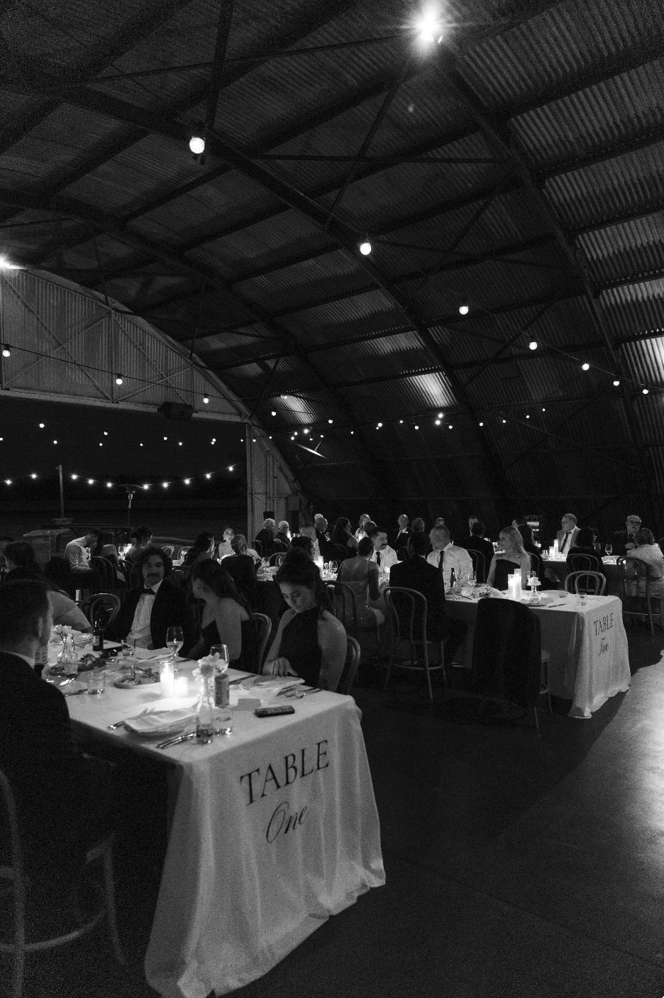 A black and white photo of a formal banquet in a large, arched hall with string lights. Guests are seated at long tables with white tablecloths, candles, and place settings. The atmosphere appears elegant and festive.