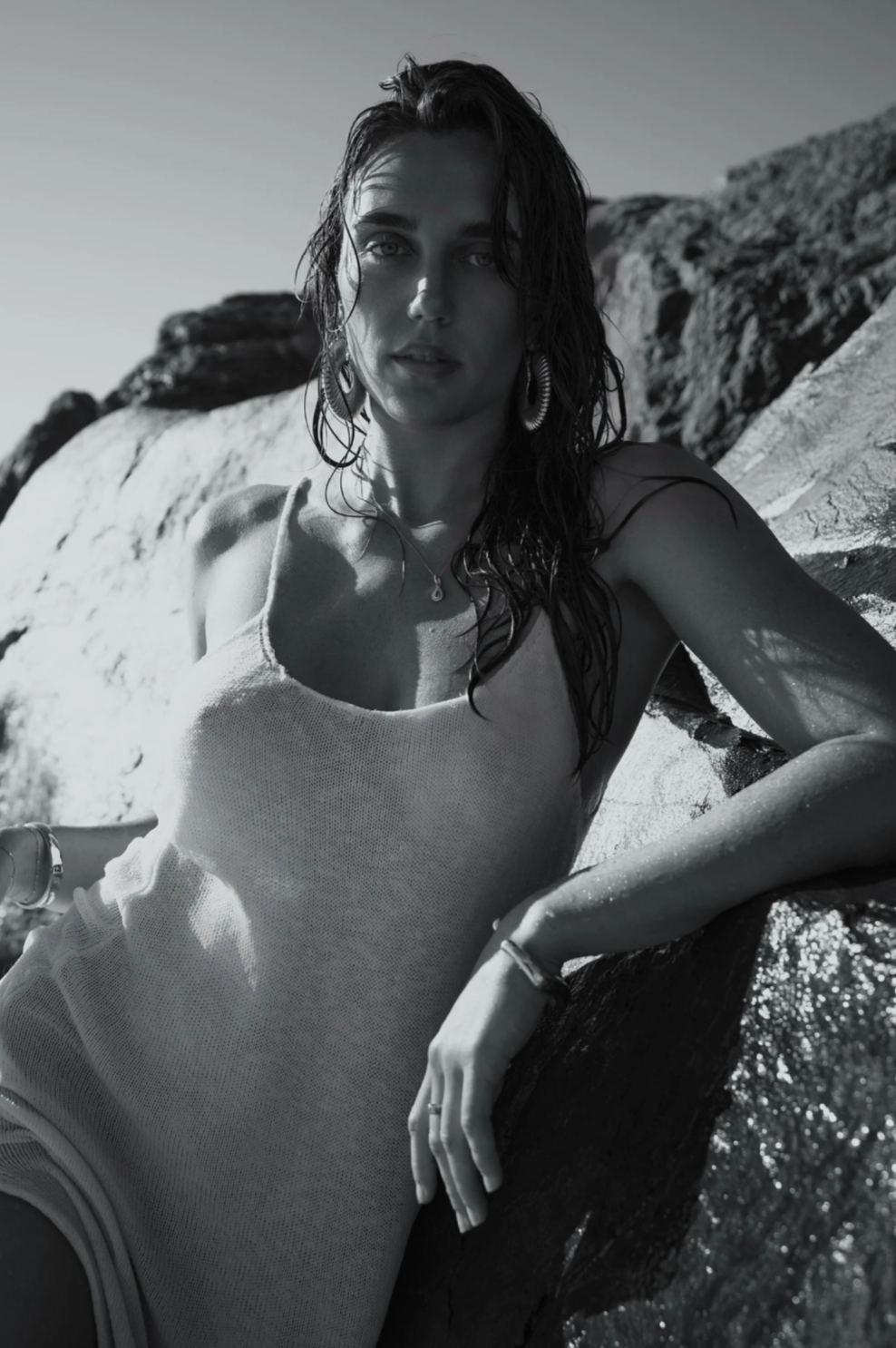 A woman with wet hair, wearing earrings and a sleeveless top, is leaning against a large rock outdoors on a sunny day.