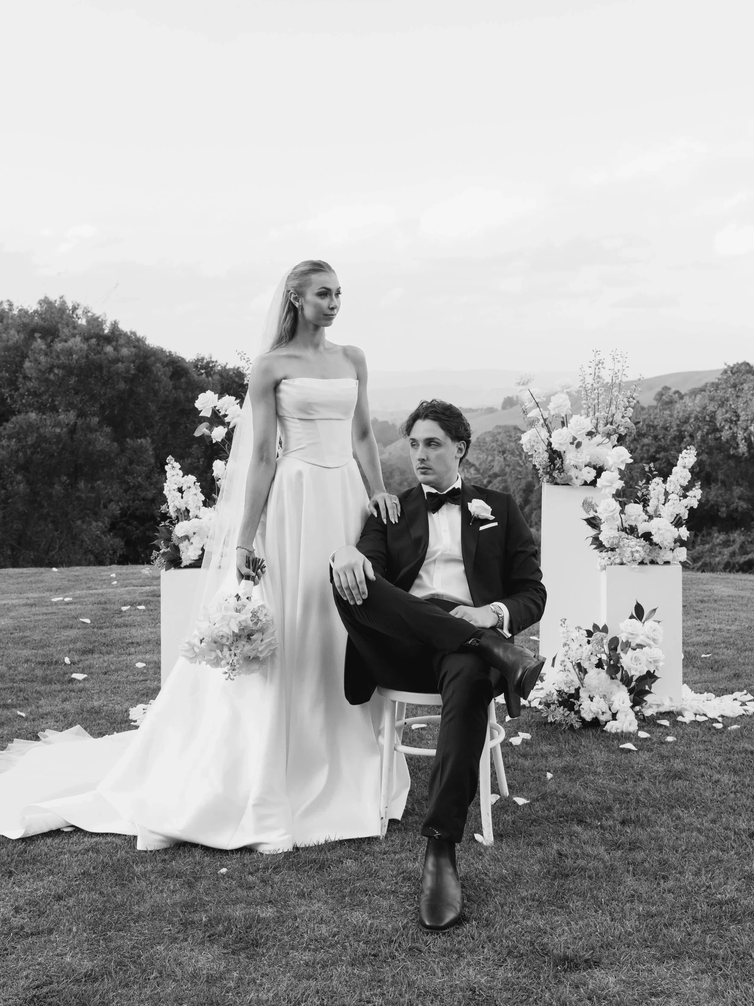 Black and white photo of a wedding couple outdoors. The woman in a strapless wedding gown with a long train, holding a bouquet, stands next to a man in a tuxedo who is sitting on a chair. The scene is decorated with floral arrangements on pedestals, and there are trees and a cloudy sky in the background.