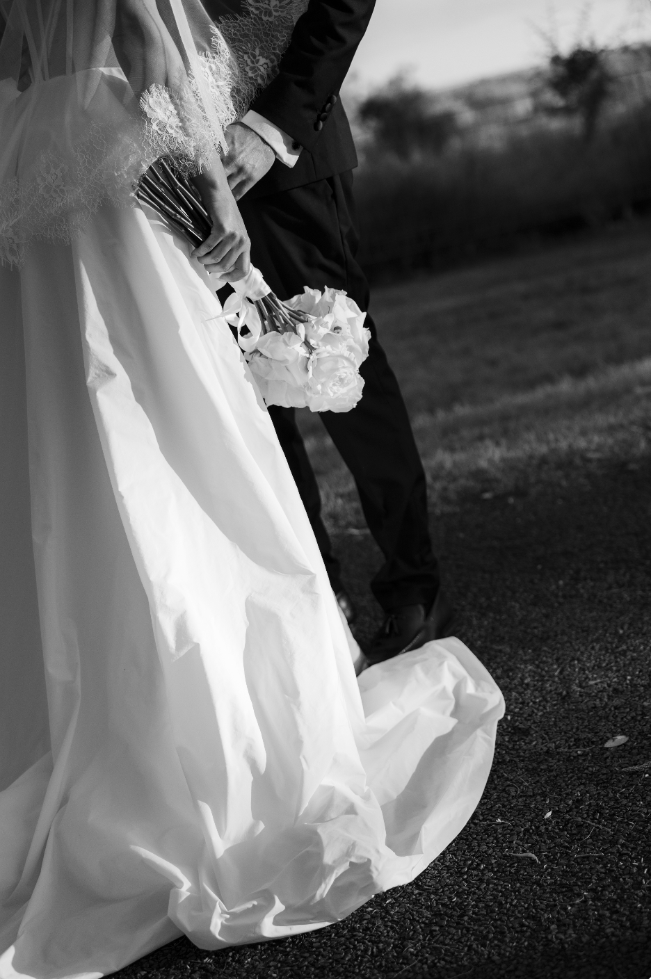 A bride and groom standing outdoors, with the bride holding a bouquet of flowers, dressed in a wedding gown and veil, and the groom in a suit.