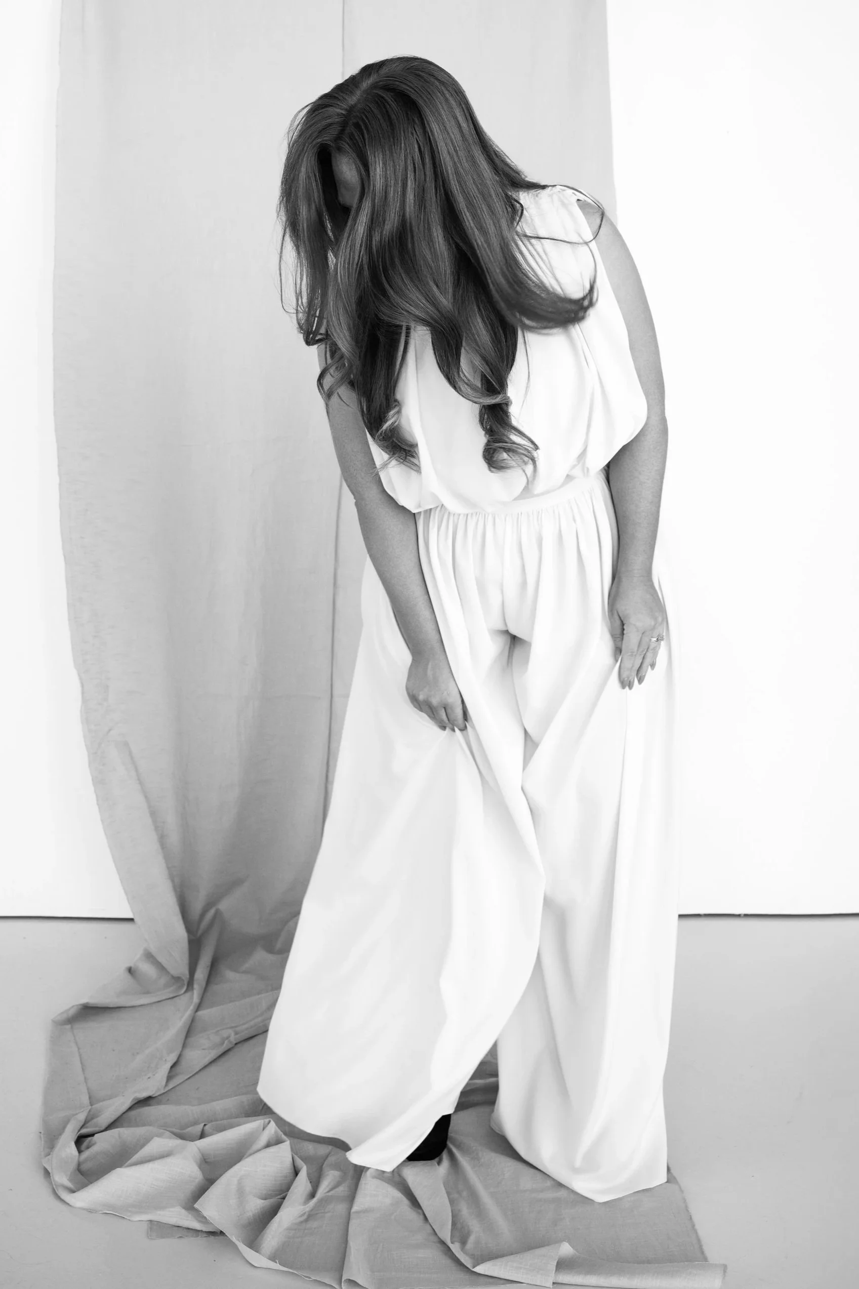 A woman with long wavy hair standing on a floor covered with crumpled fabric, leaning her head downward and wearing a loose white outfit, in front of a plain white wall.